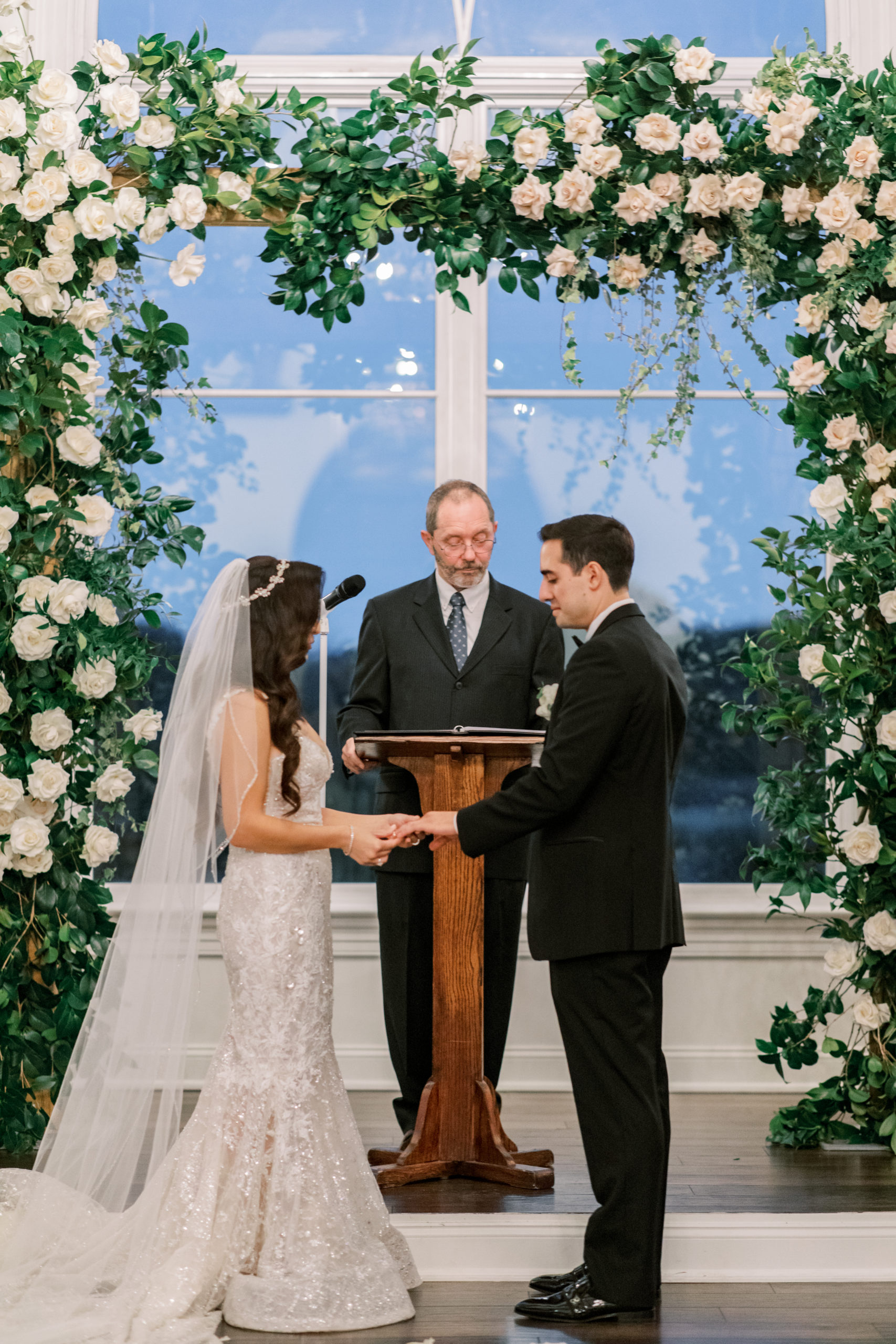 bride and groom exchange rings during wedding ceremony under rose filled square floral arch