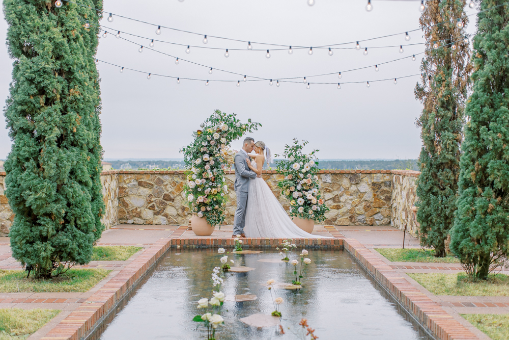 bride and groom kiss in front of reflecting pool with large floral arch