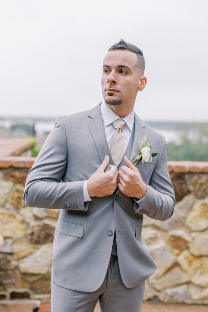 groom in grey suit stands in front of stone wall Bella Collina Wedding