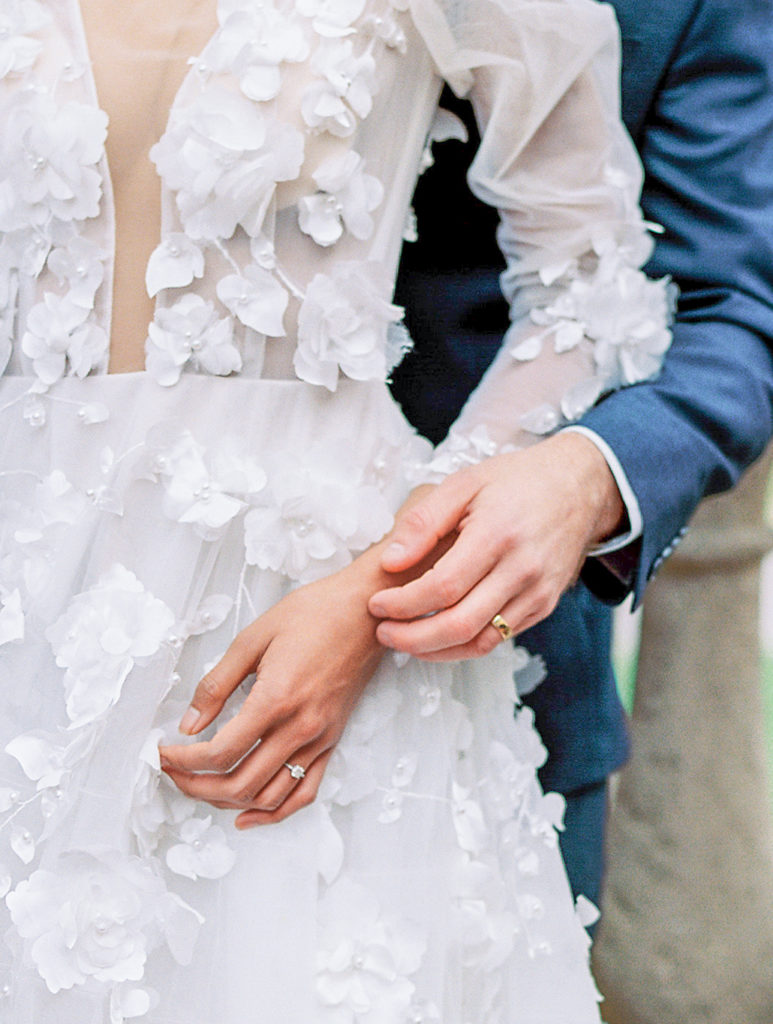 clise up of bride and groom's hands with blue suit and floral applique dress Bella Collina Wedding Photography