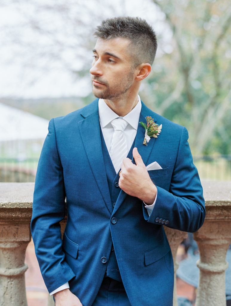 groom in blue suit stands on stone terrace