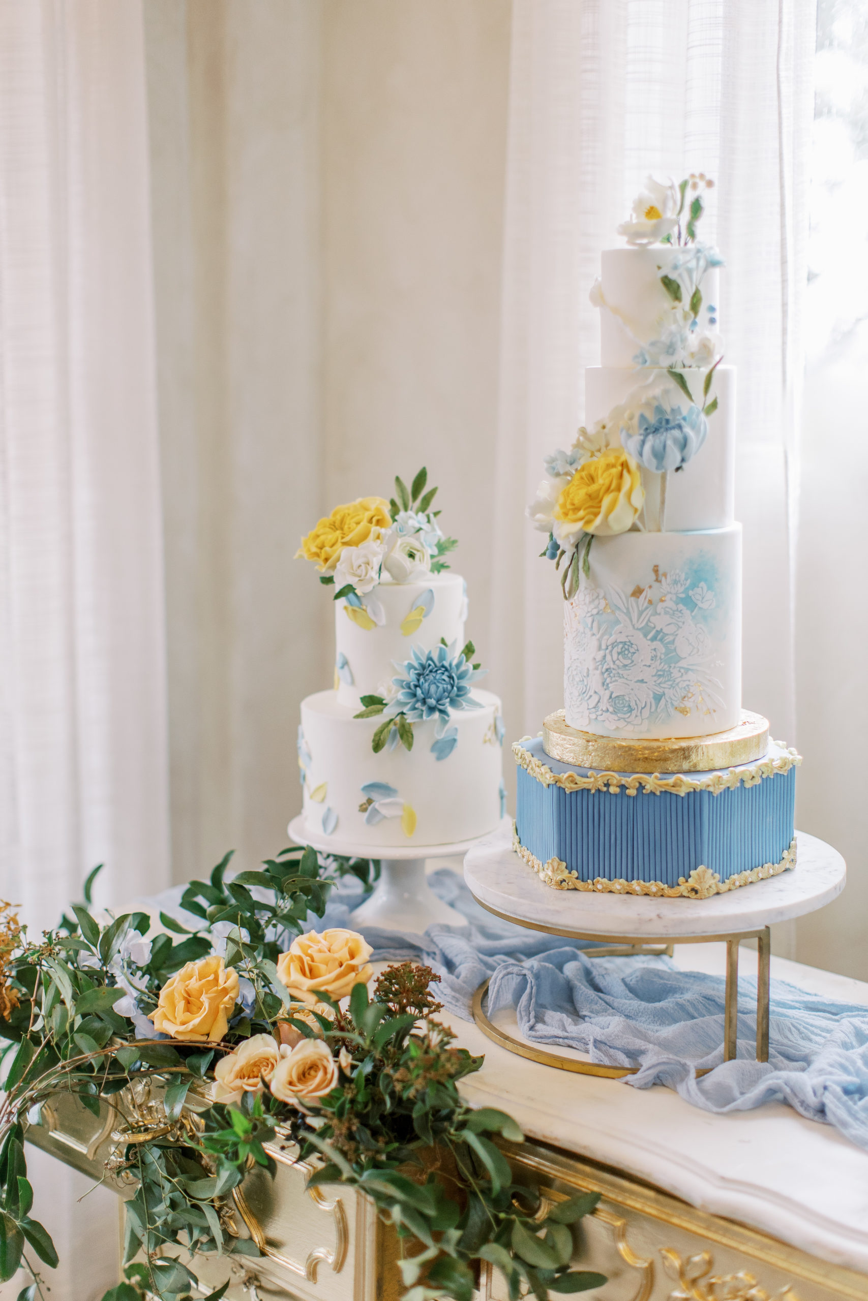 Two blue, white, and gold wedding cakes on antique dresser with golden flowers
