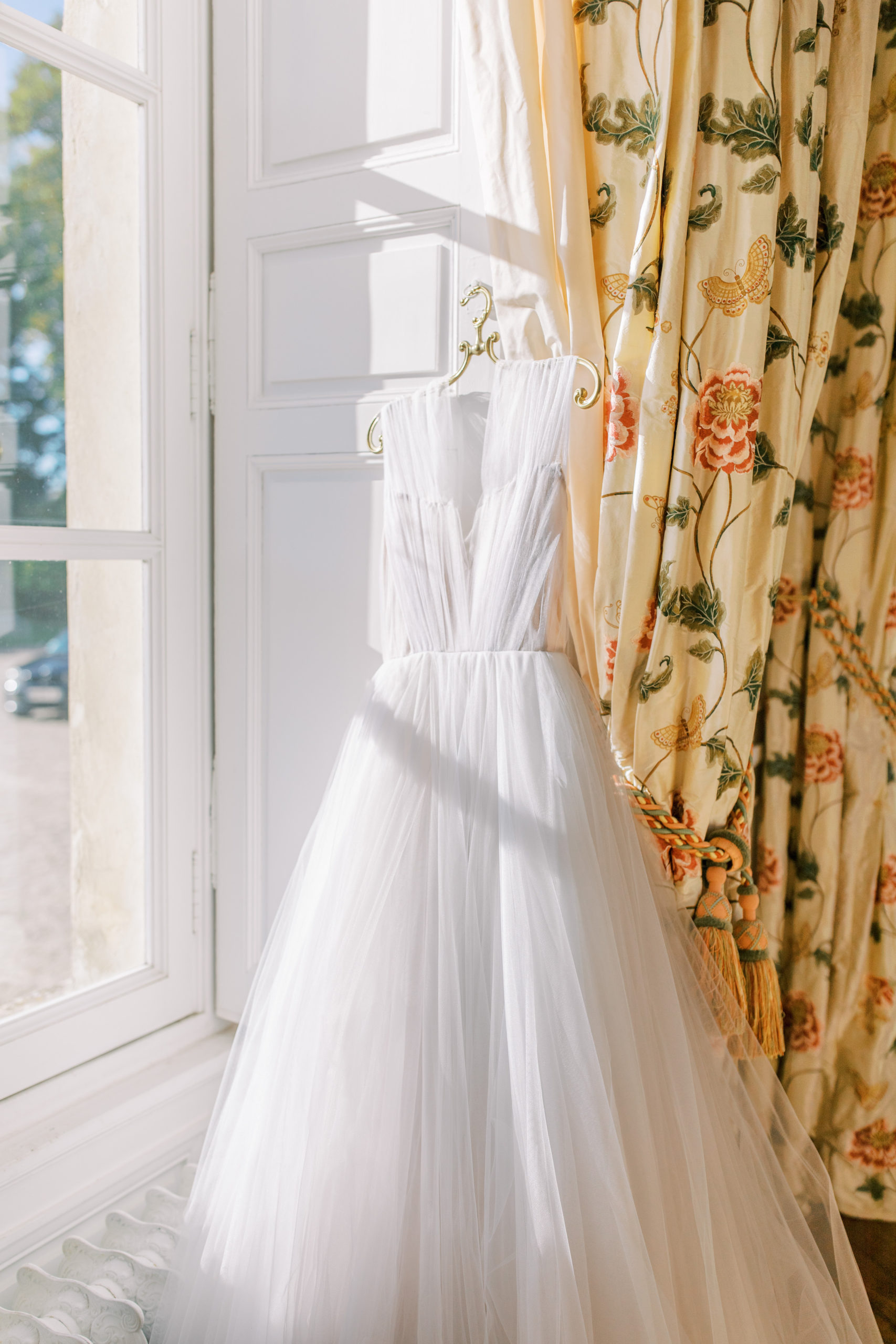 Wedding Dress hangs in the window with sunlight and gold curtains