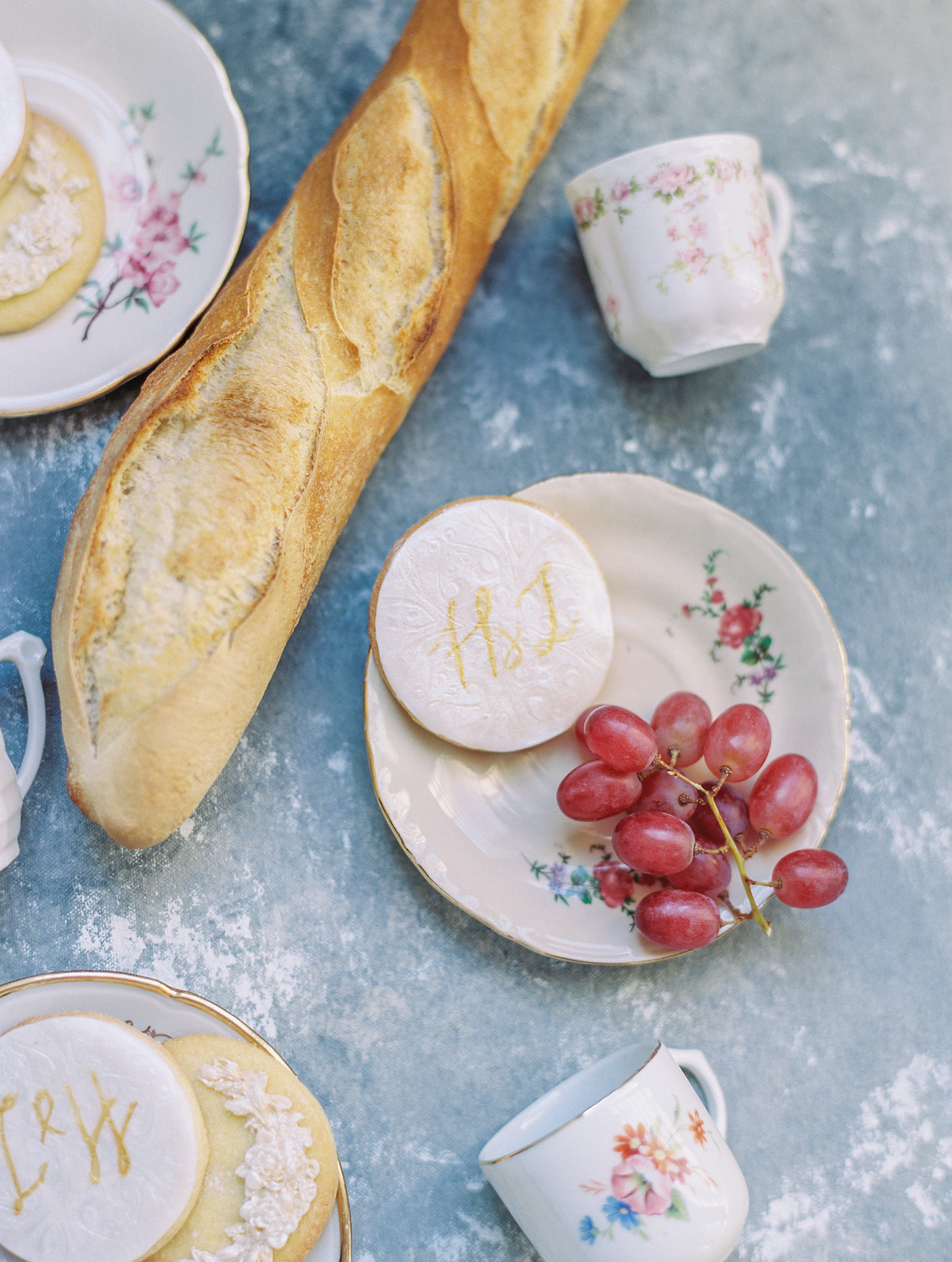 Up close of baguette, grapes, and couple initialed cookies with pink, cream, red, and green tea cups and plates 