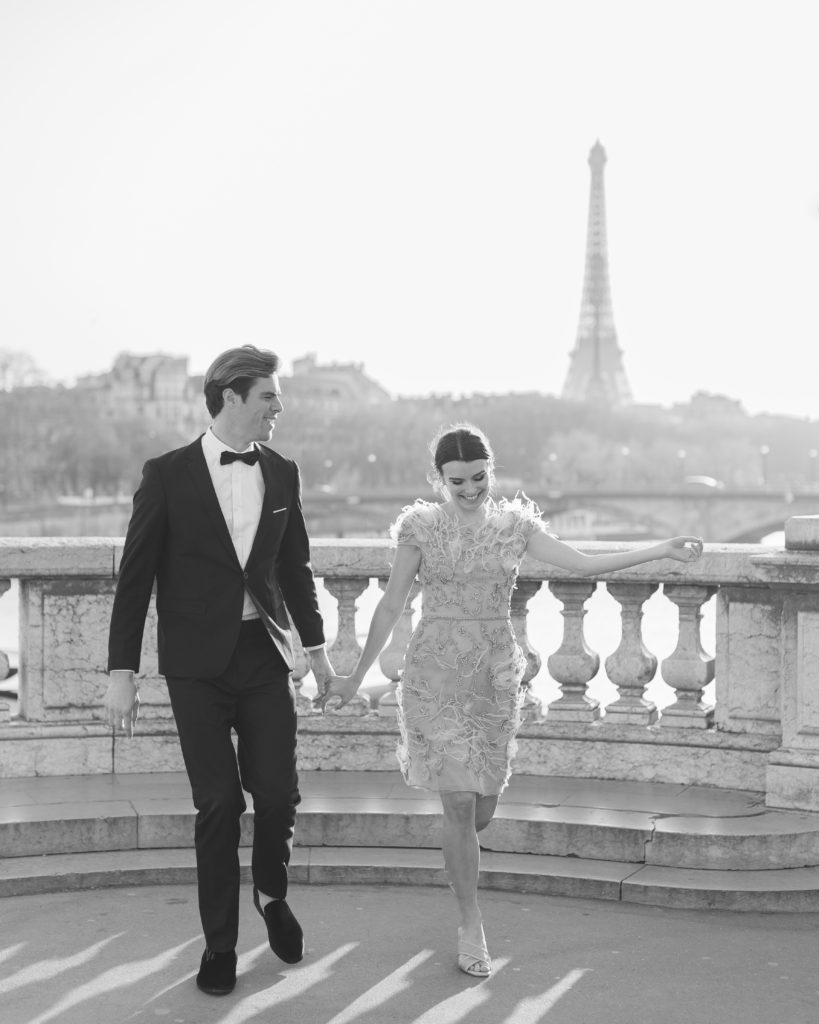 Couple holding hands smiling walking with Eiffel Tower in background for Sunset Paris Engagement 