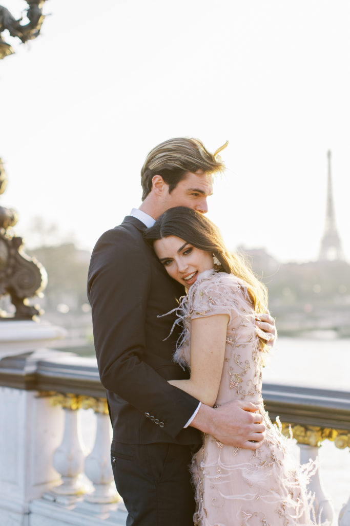 Couple embracing into each other on bridge with Eiffel Tower in background for Sunset Paris Engagement 