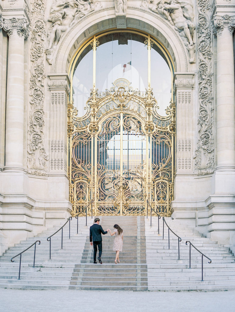 Couple hold hands walking away on stairs in front of golden gates for Sunset Paris Engagement 