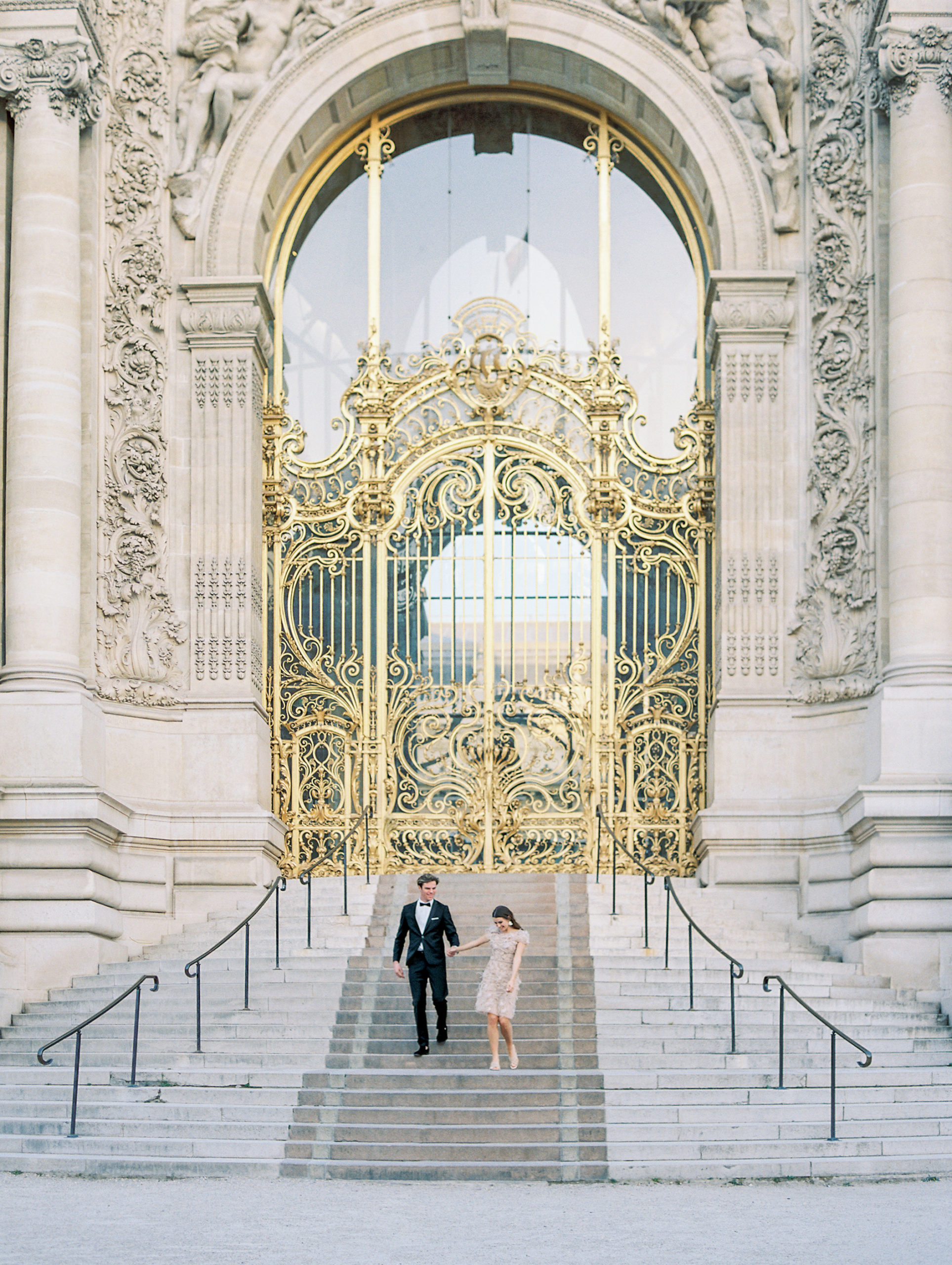 Couple holding hands walking down stairs in front of golden gates for Sunset Paris Engagement  