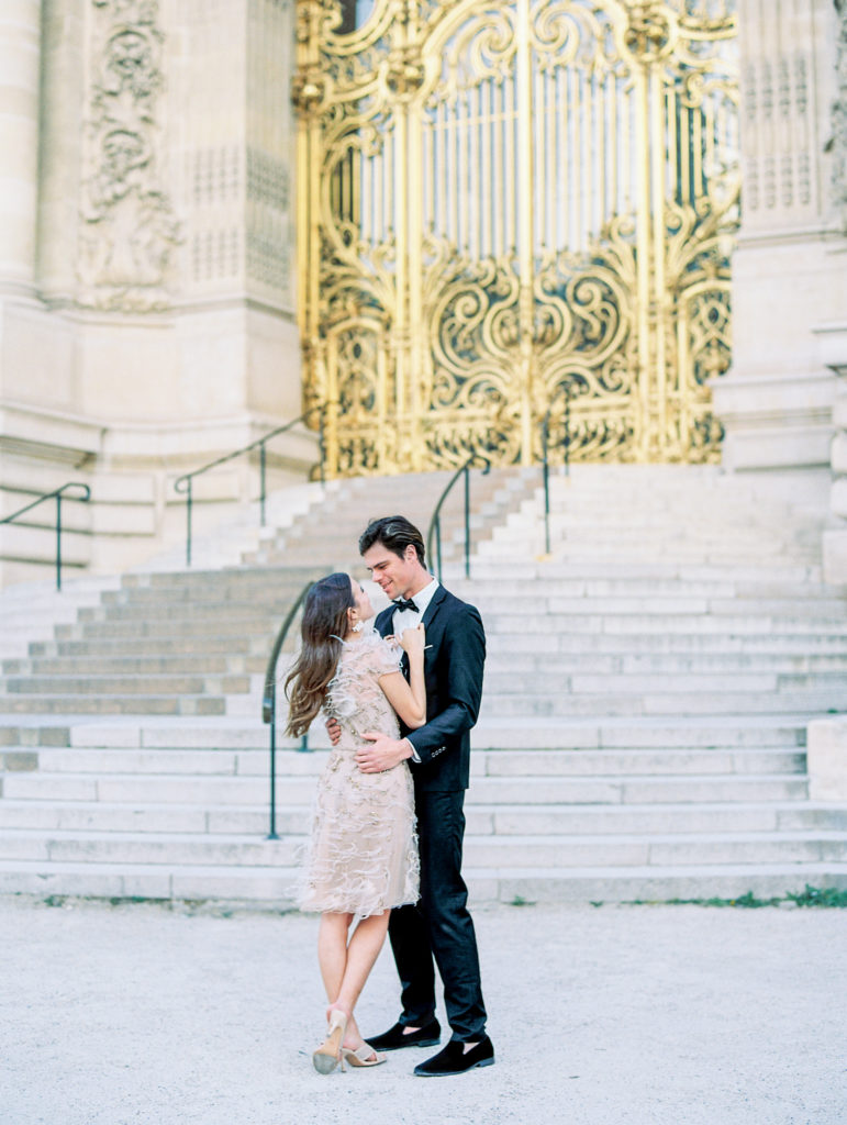 Couple embraces in front of stairs and golden gates for Sunset Paris Engagement 