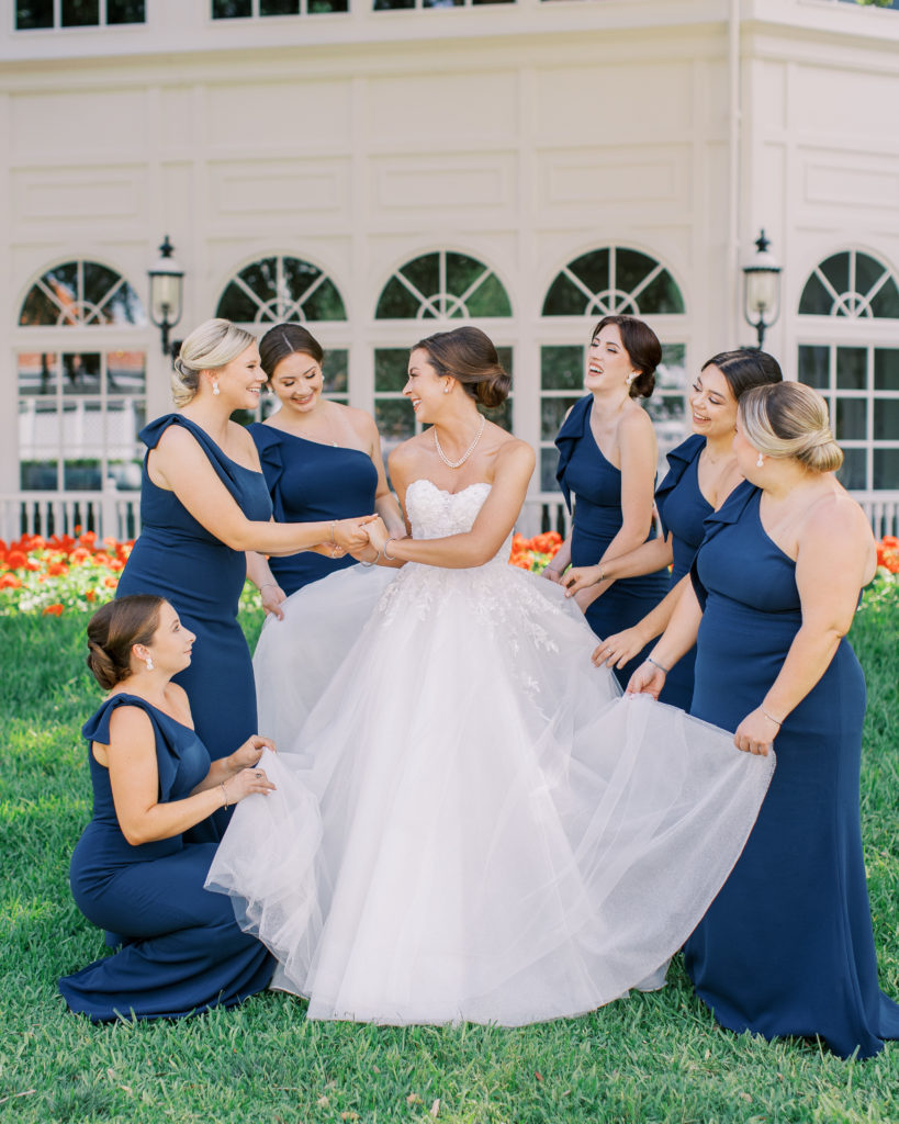 Bridesmaids stand around bride holding up dress and smile and laugh 