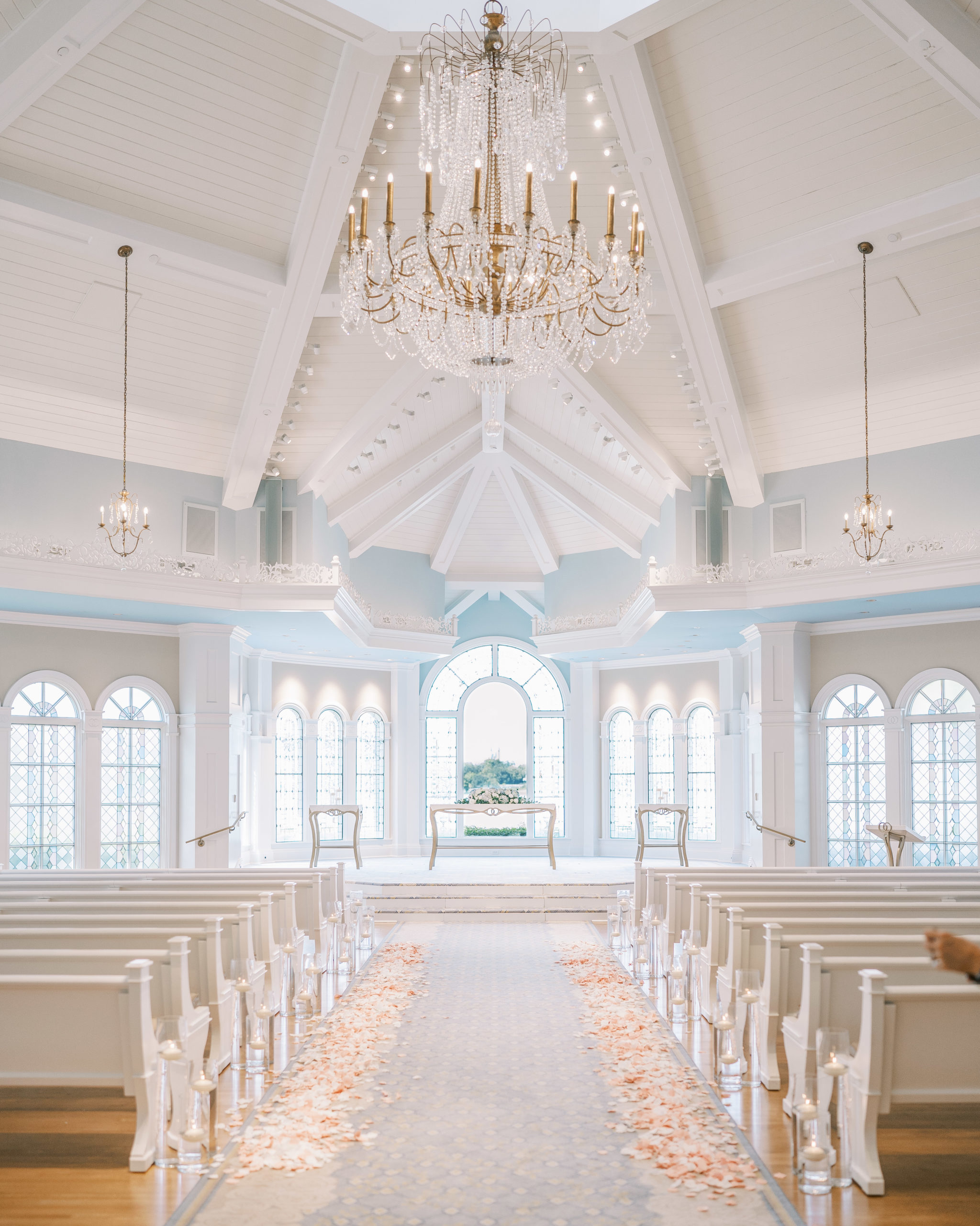 Light blue and white wedding ceremony with chandelier and white pews with pink and white petals lining seats 
