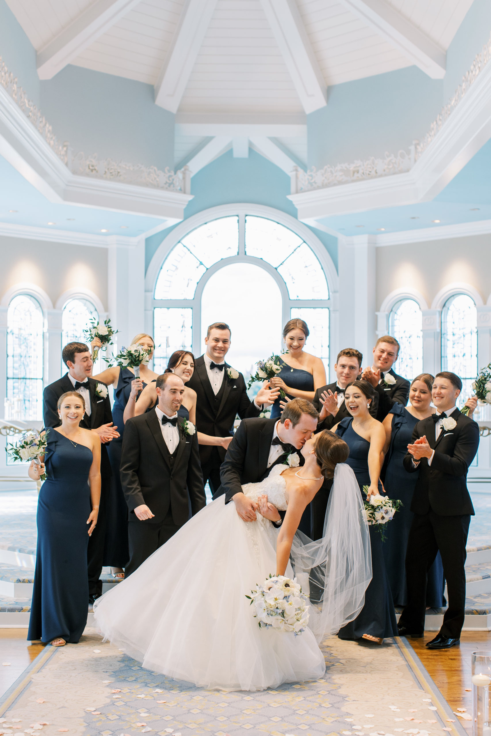 Bride and groom kiss in blue and white pavilion as wedding party surrounds and smiles 