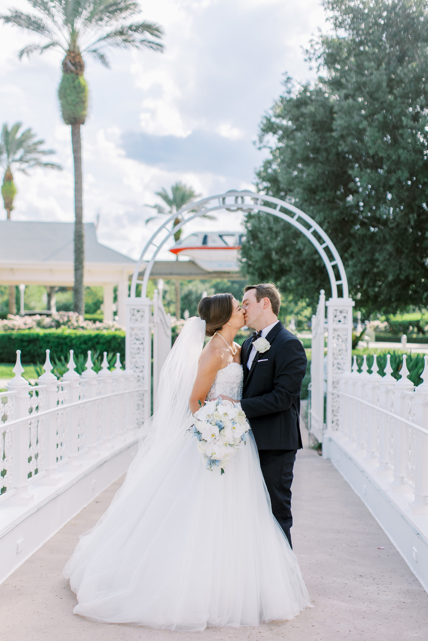 Bride and groom kiss on white bridge under arch with palm tree at a Romantic and Timeless Grand Floridian Wedding 