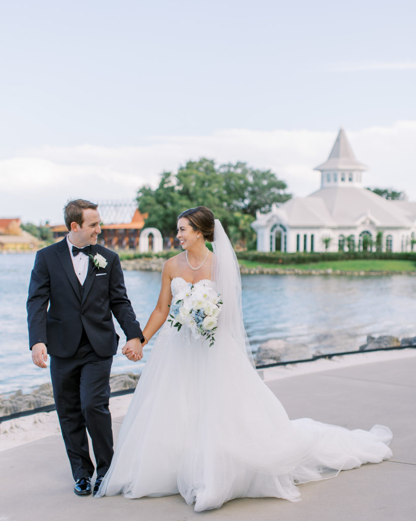 Bride and groom hold hands walking alongside lake at a Romantic and Timeless Grand Floridian Wedding 