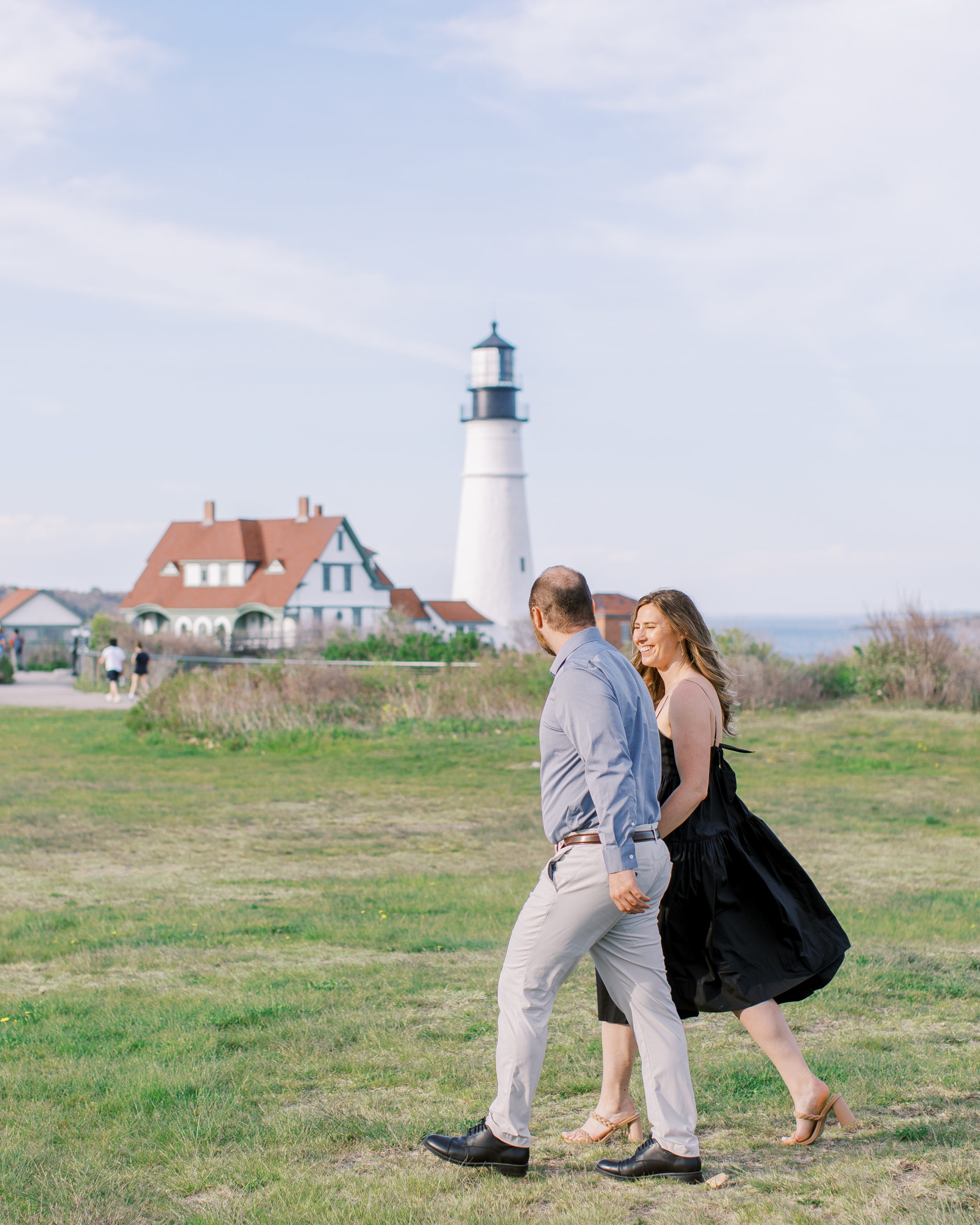 Couple holding hands walking in field with lighthouse in Maine 