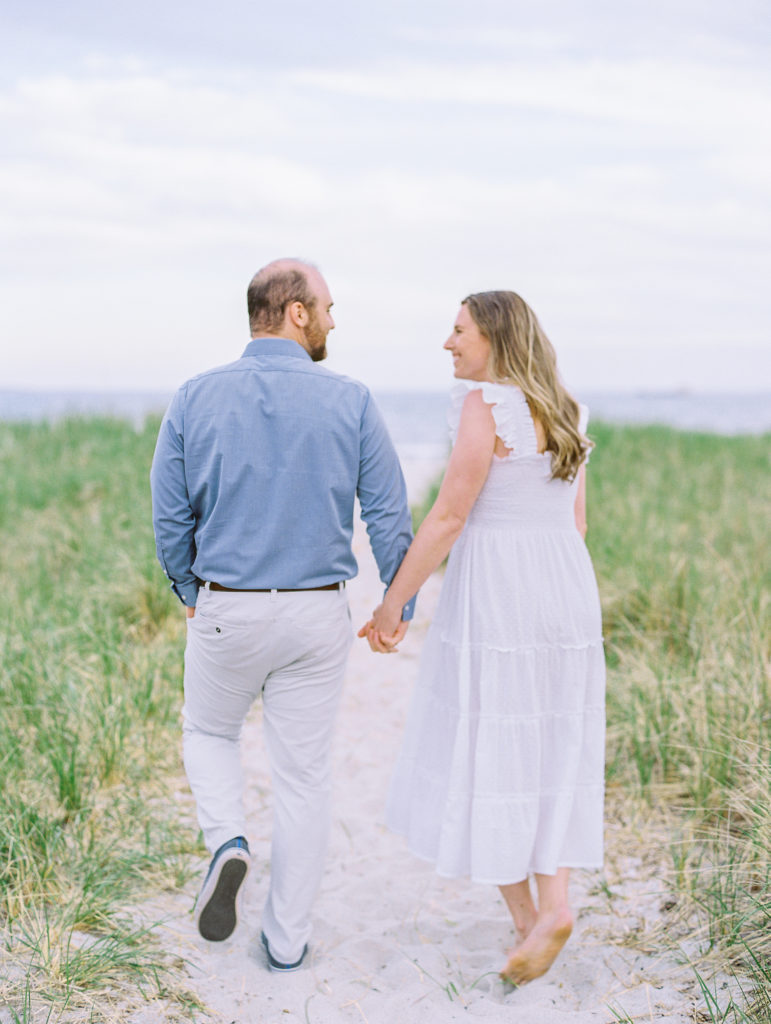 Couple holding hands walking away towards the beach smiling for Coastal Maine Engagement Session 
