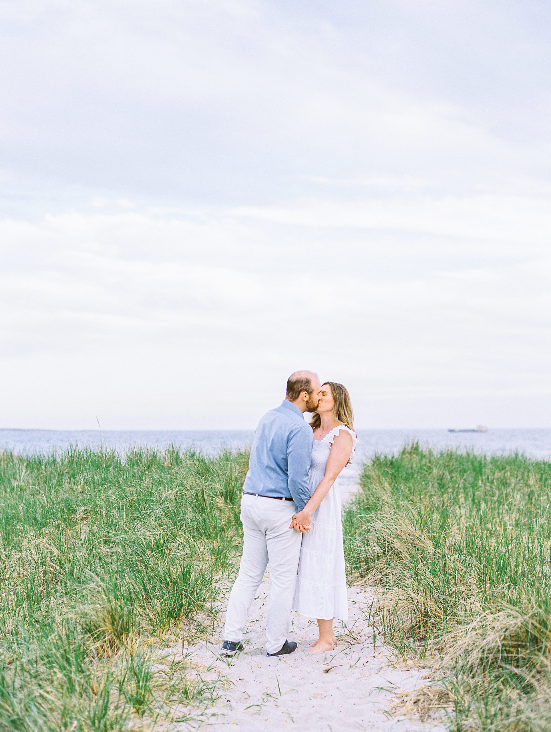 Couple holds hands and kiss on beach pathway on dune towards the ocean for Coastal Maine Engagement Session 