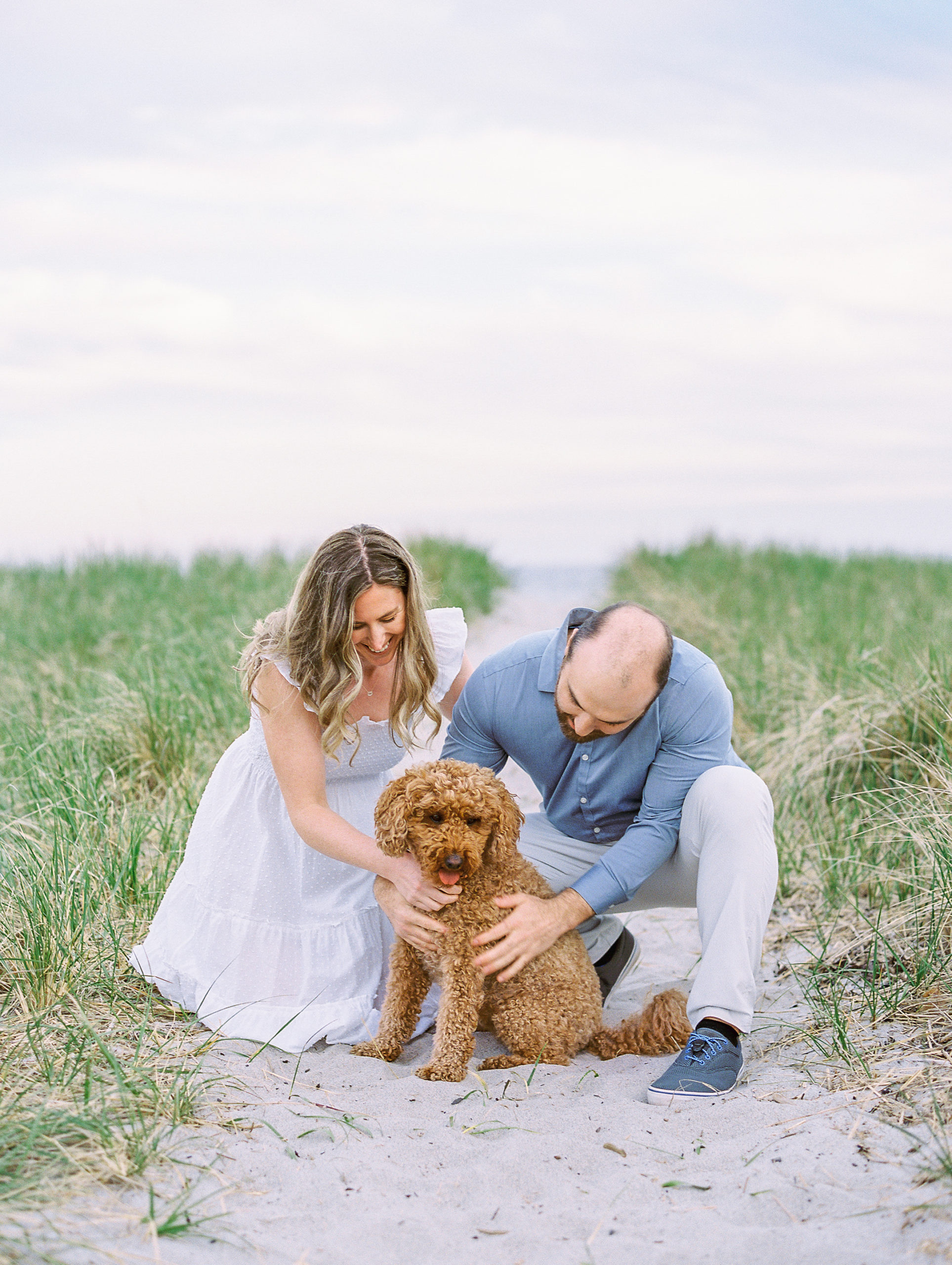 Couple smile and embrace dog on beach pathway for Coastal Maine Engagement Session 