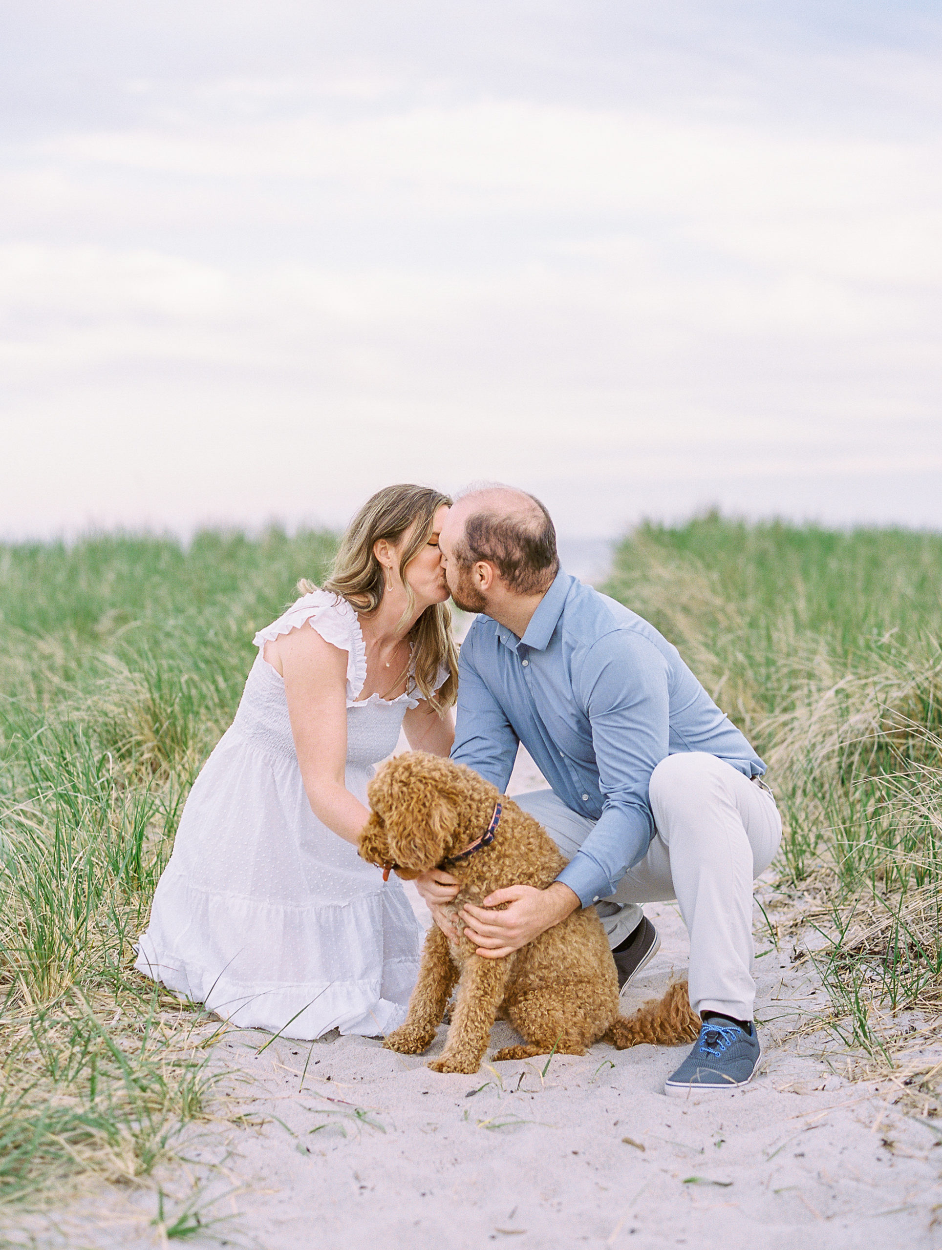 Couple embrace dog and kiss on beach pathway for Coastal Maine Engagement Session 