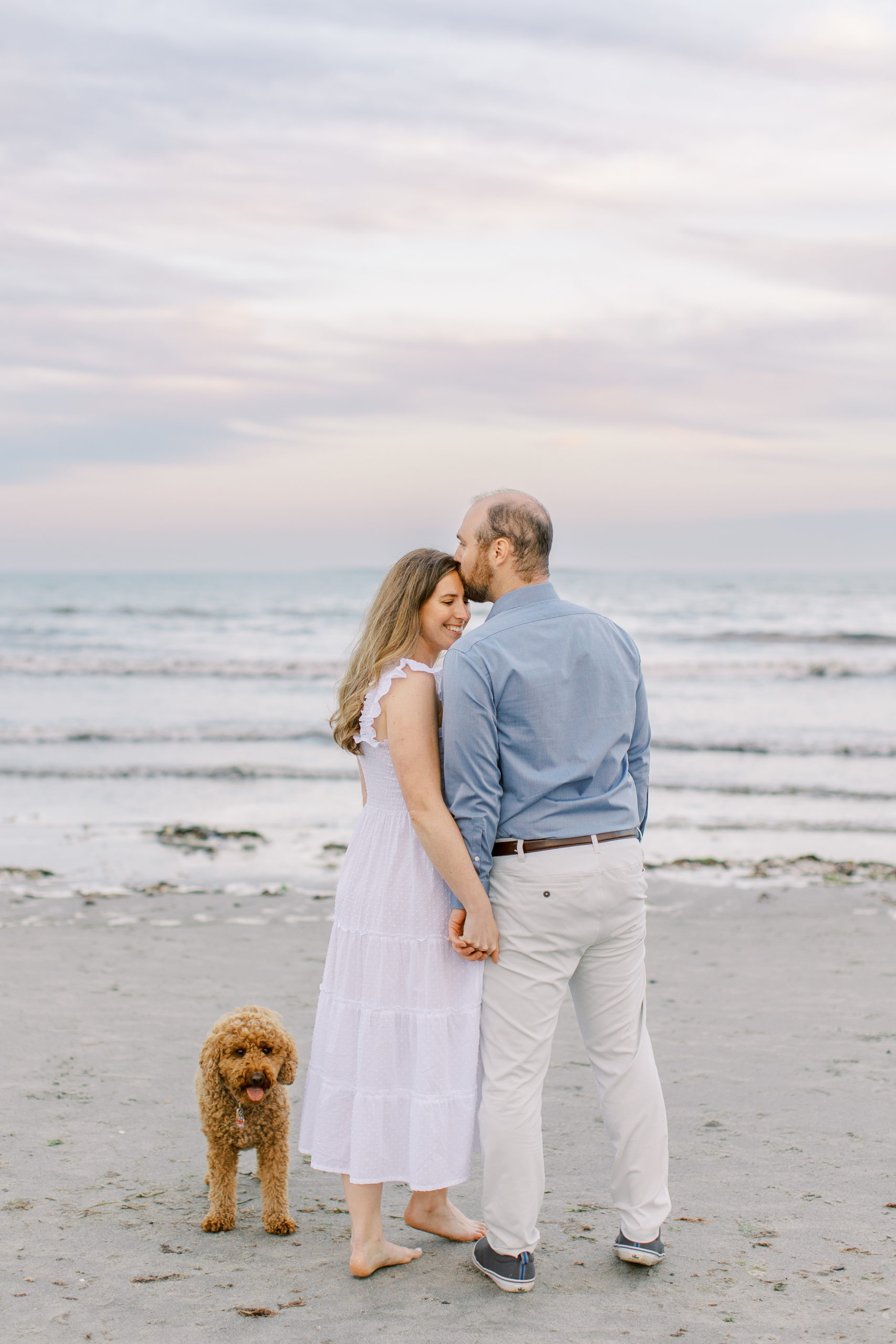 Couple holds hand with a kiss on forehead and dog by the side on the beach for Coastal Maine Engagement Session 