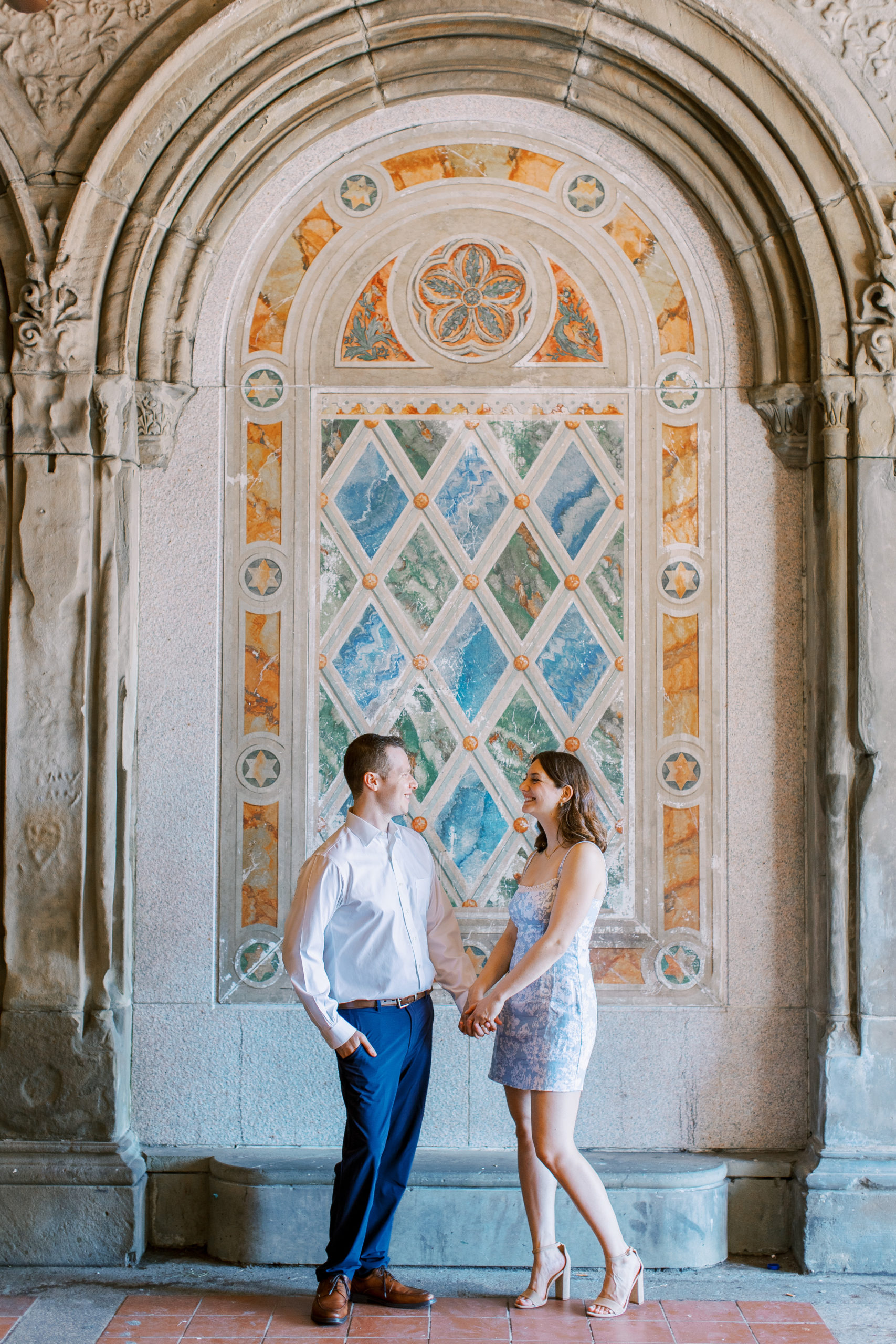Couple holds hand and smiles in front of blue, green, and orange tiles inside terrace arcade for Central Park Engagement Photos