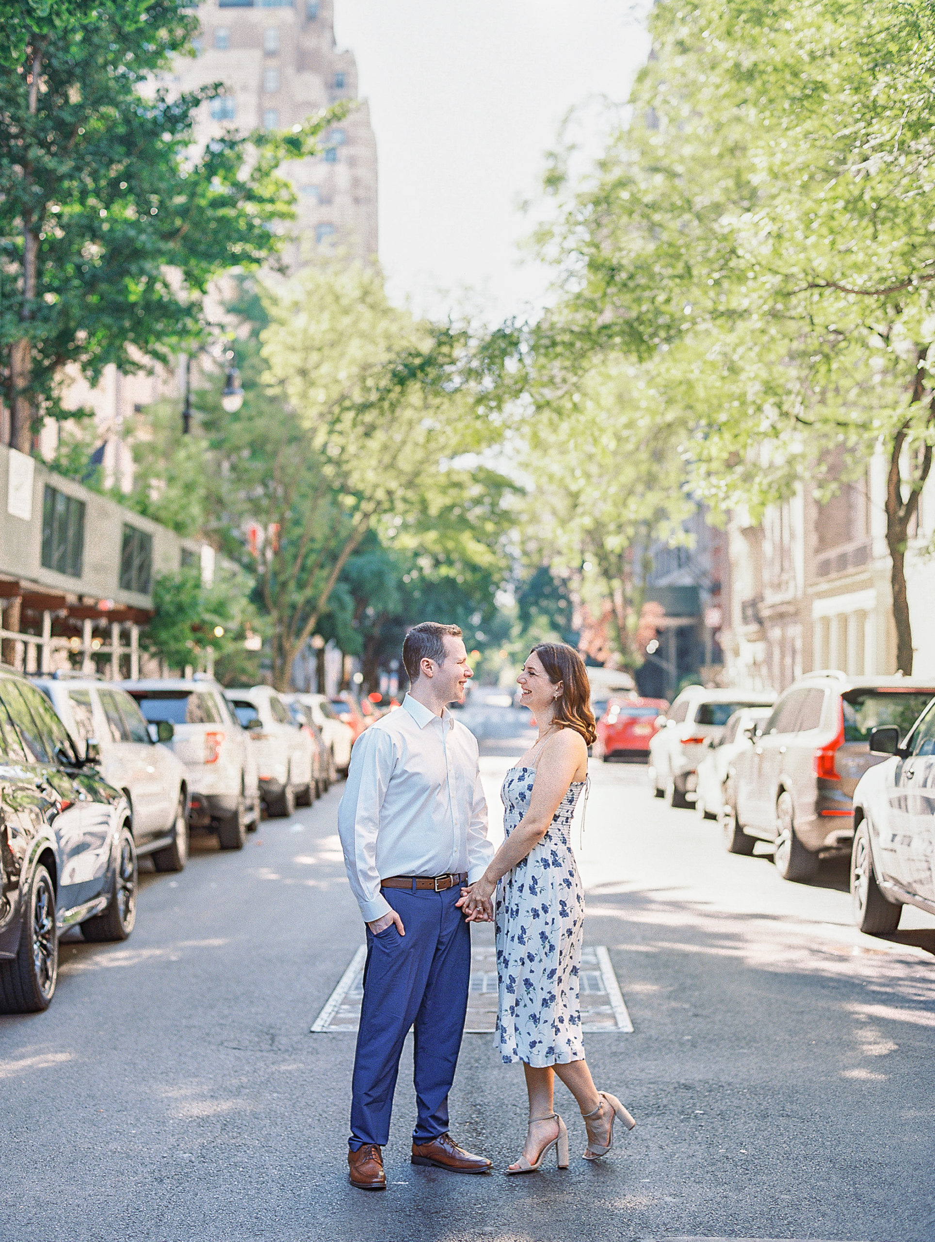 Couple hold hands and smile at each other in middle of tree lined road in New York City for Central Park Engagement Photos