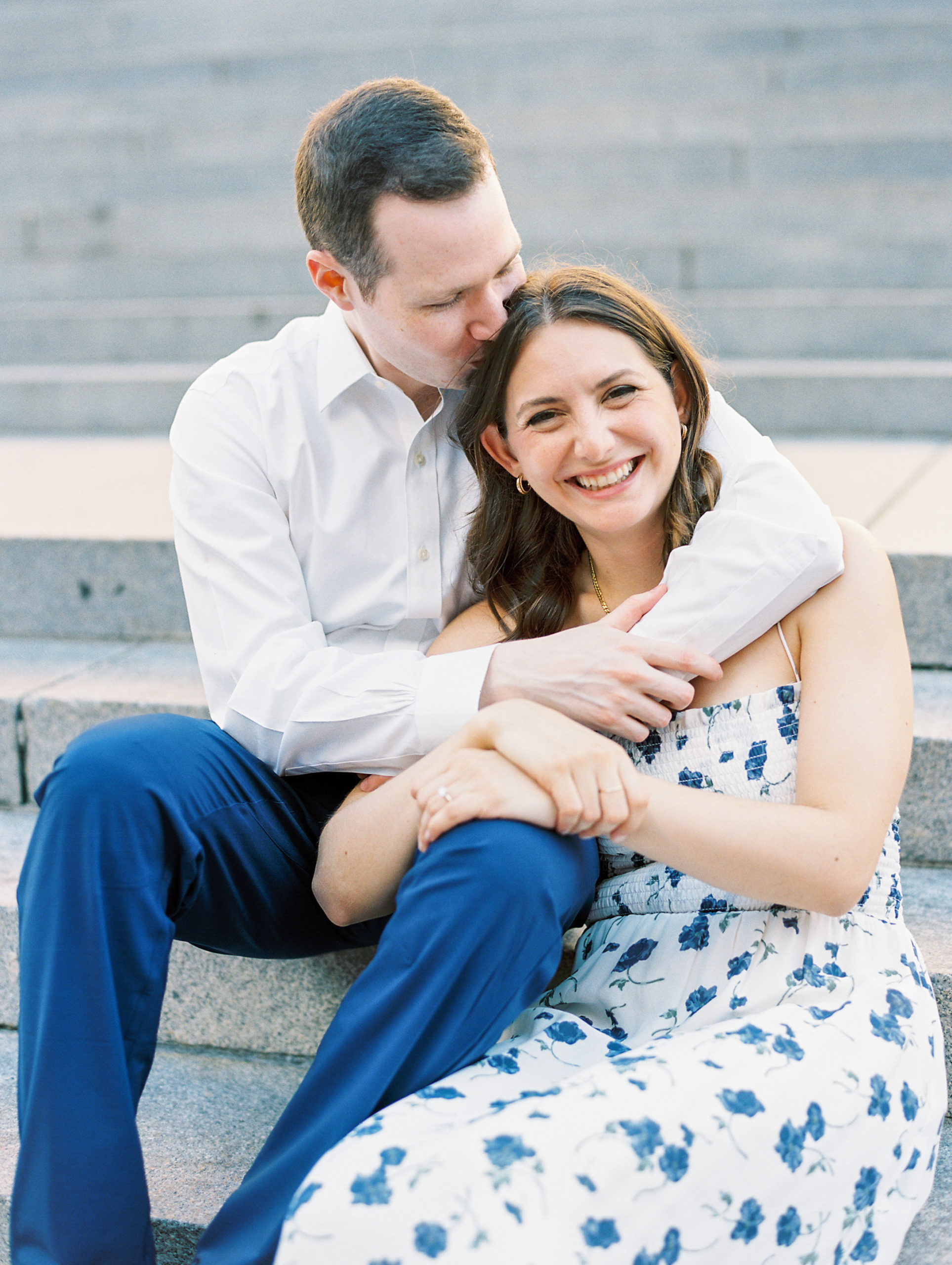 Fiancé embraces and kisses head of partner as she smiles on stone stairs of museum for Central Park Engagement Photos