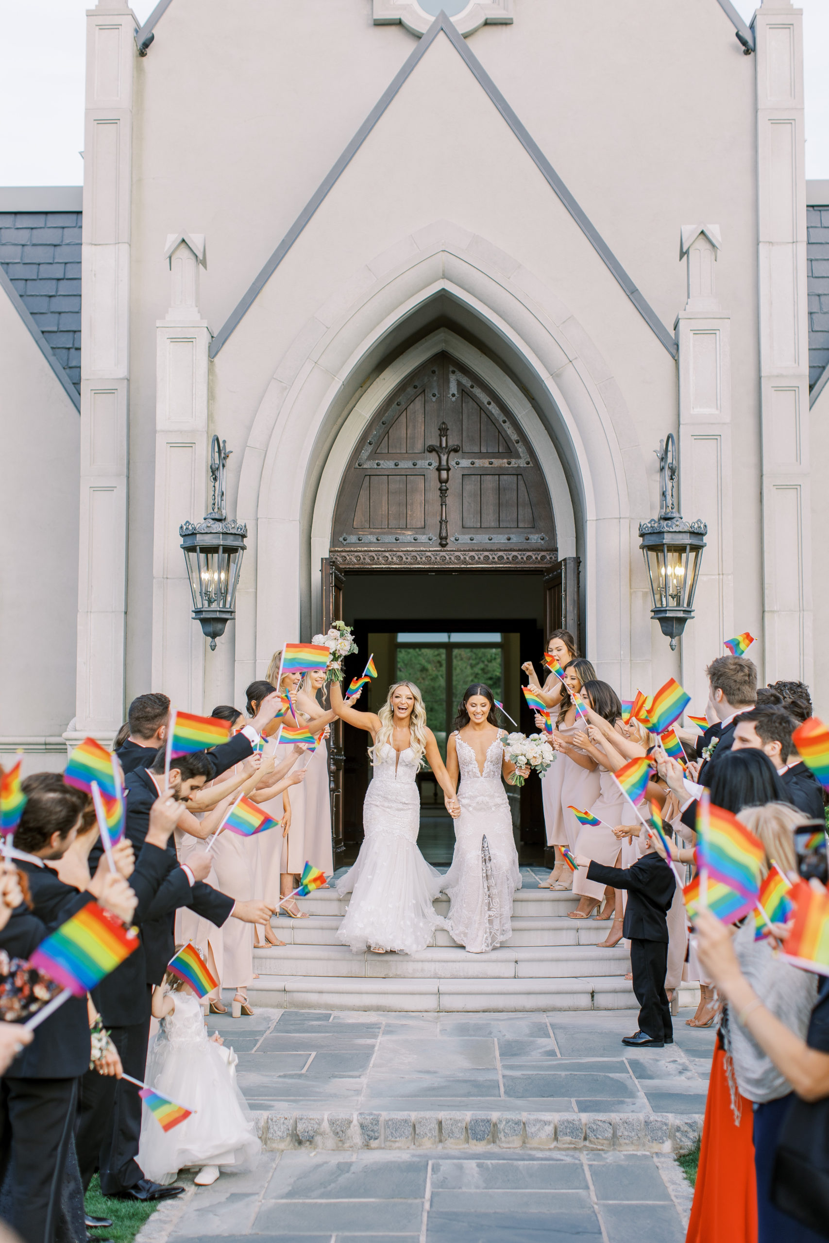 Brides hold hands with bridal party and friends holding pride flags at a Romantic Park Chateau Wedding Photography