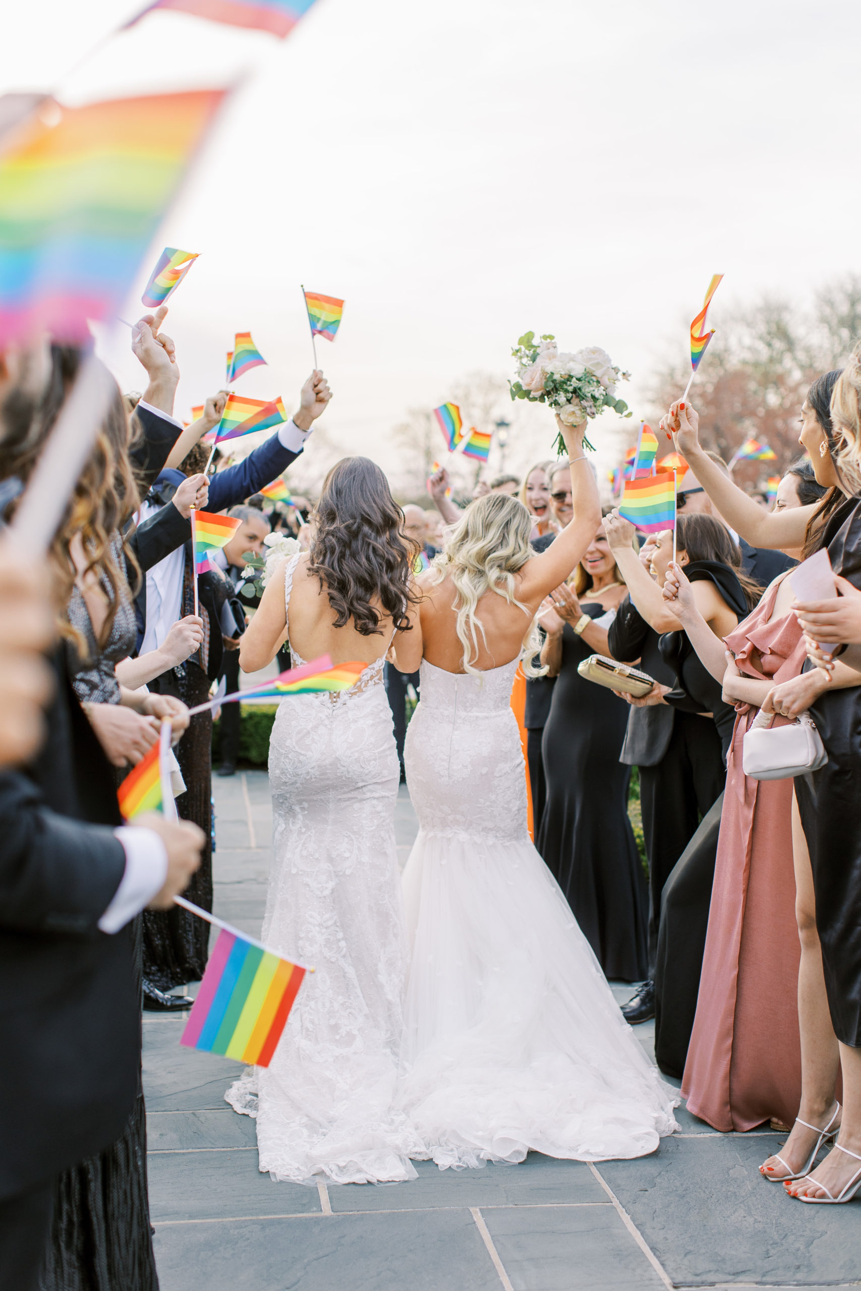 Brides hold bouquets after being married with friends and family holding pride flags at a Romantic Park Chateau Wedding Photography