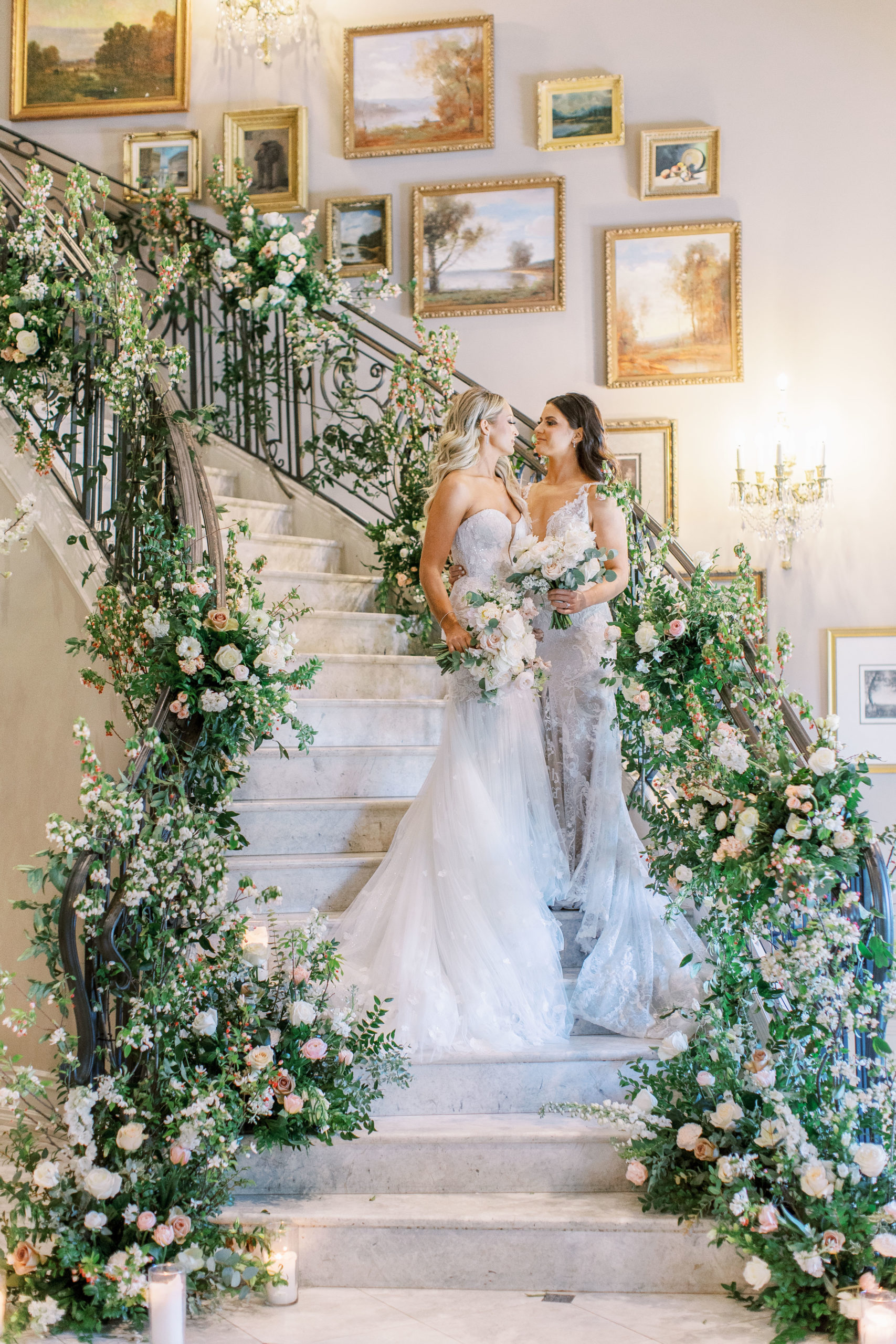 Brides embrace smiling on grand staircase with roses on banisters for a Romantic Park Chateau Wedding Photography
