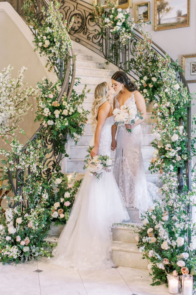 Brides kiss on grand stair case with white and pink roses on banister 
