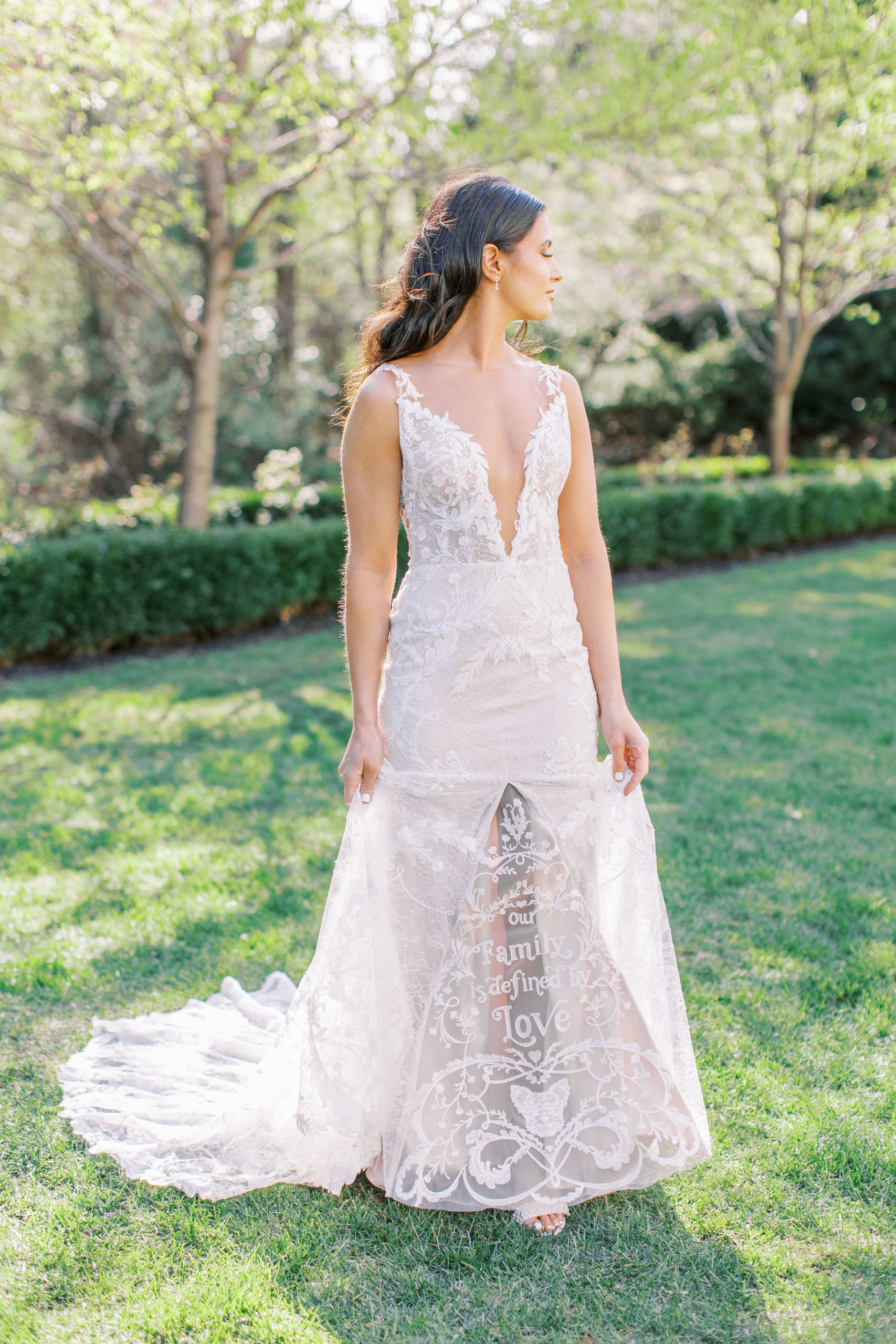 Bride holds out dress that says "Our Family is defined by Love" in garden for a Romantic Park Chateau Wedding Photography