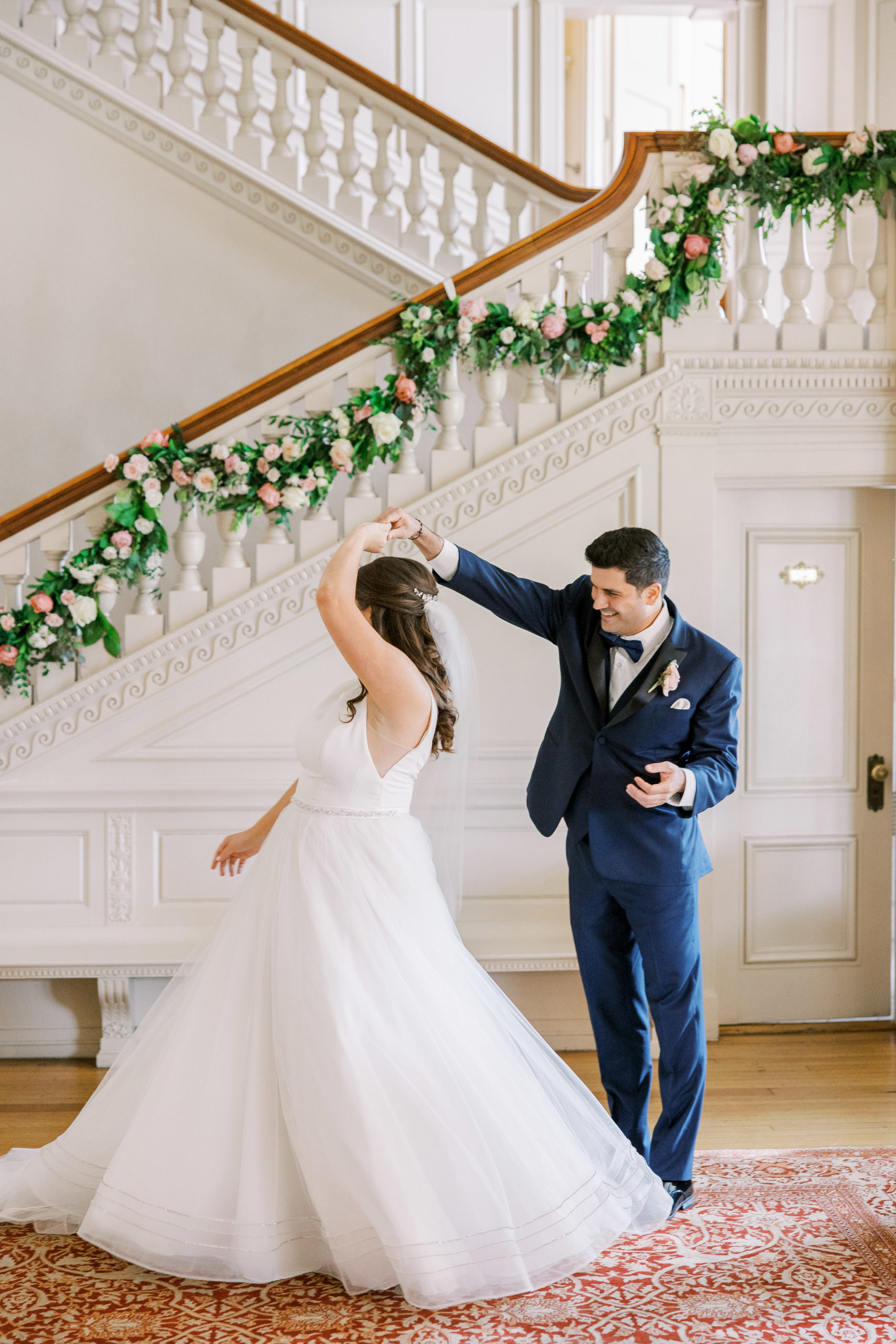 Groom spins bride around and smiles in great hall with grand staircase and rose garland 