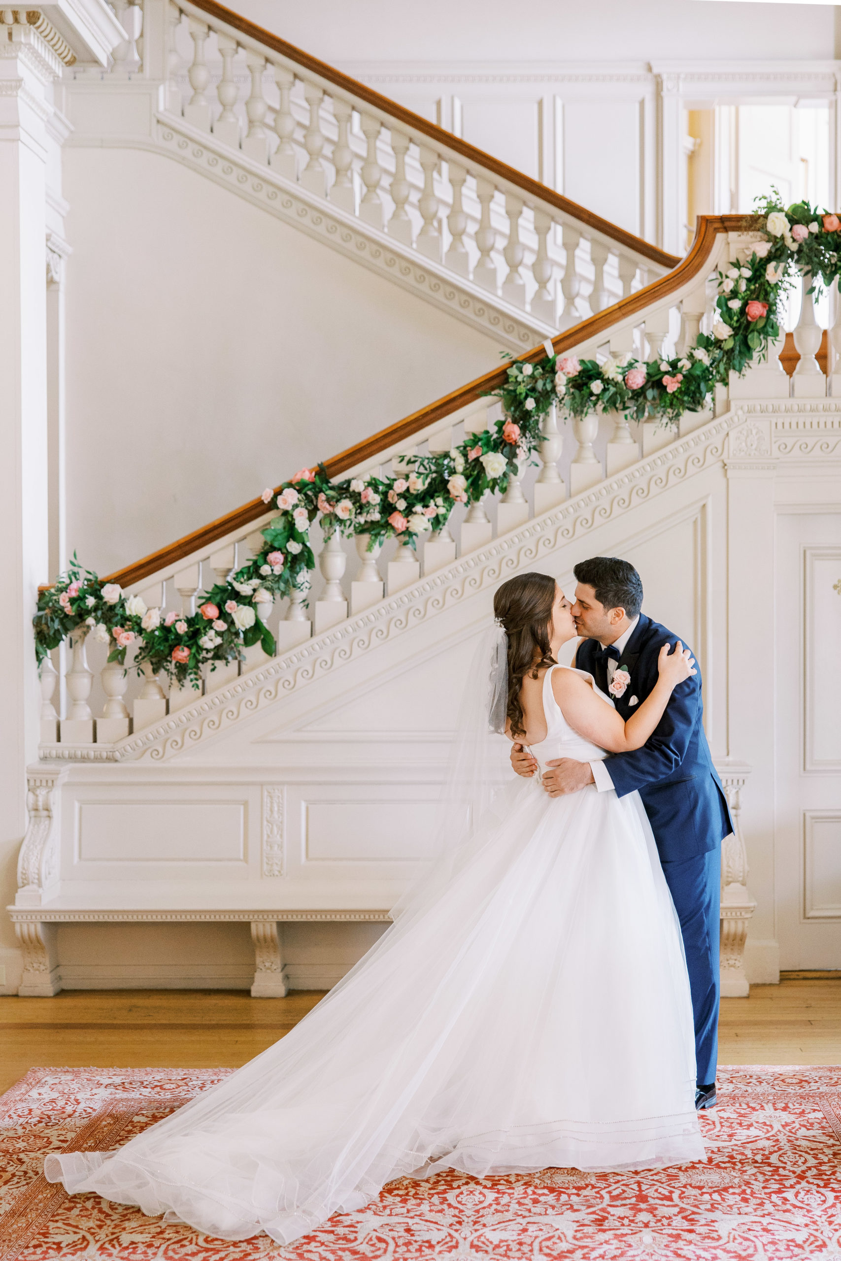 Bride and groom embrace and share a kiss in front of staircase with rose garland for Cairnwood Estate Wedding Photography