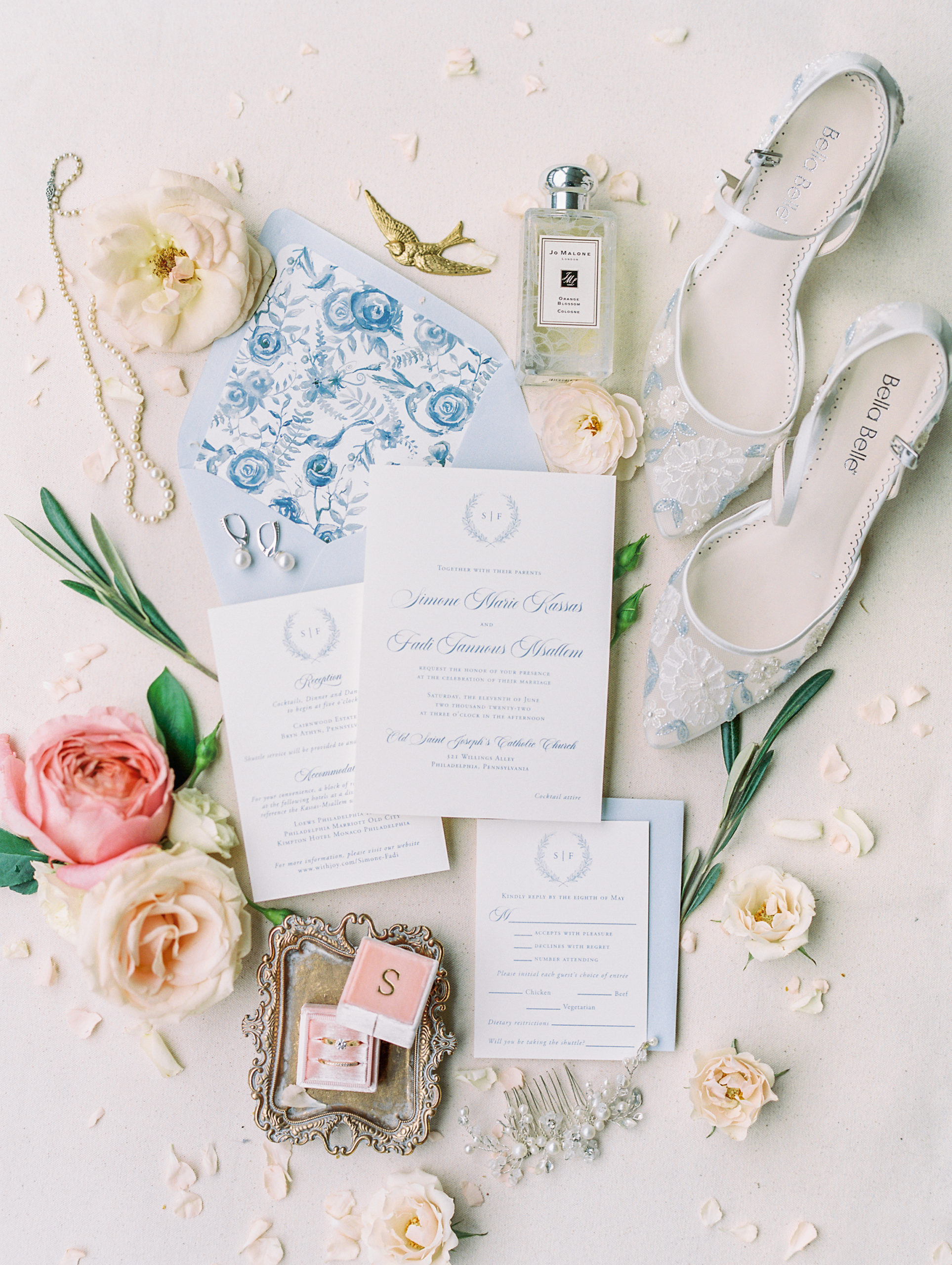 Light blue wedding invitations with delicate calligraphy, pink and cream roses, jewelry, floral white and blue heels, perfume, and pink ring box, and olive leaves 