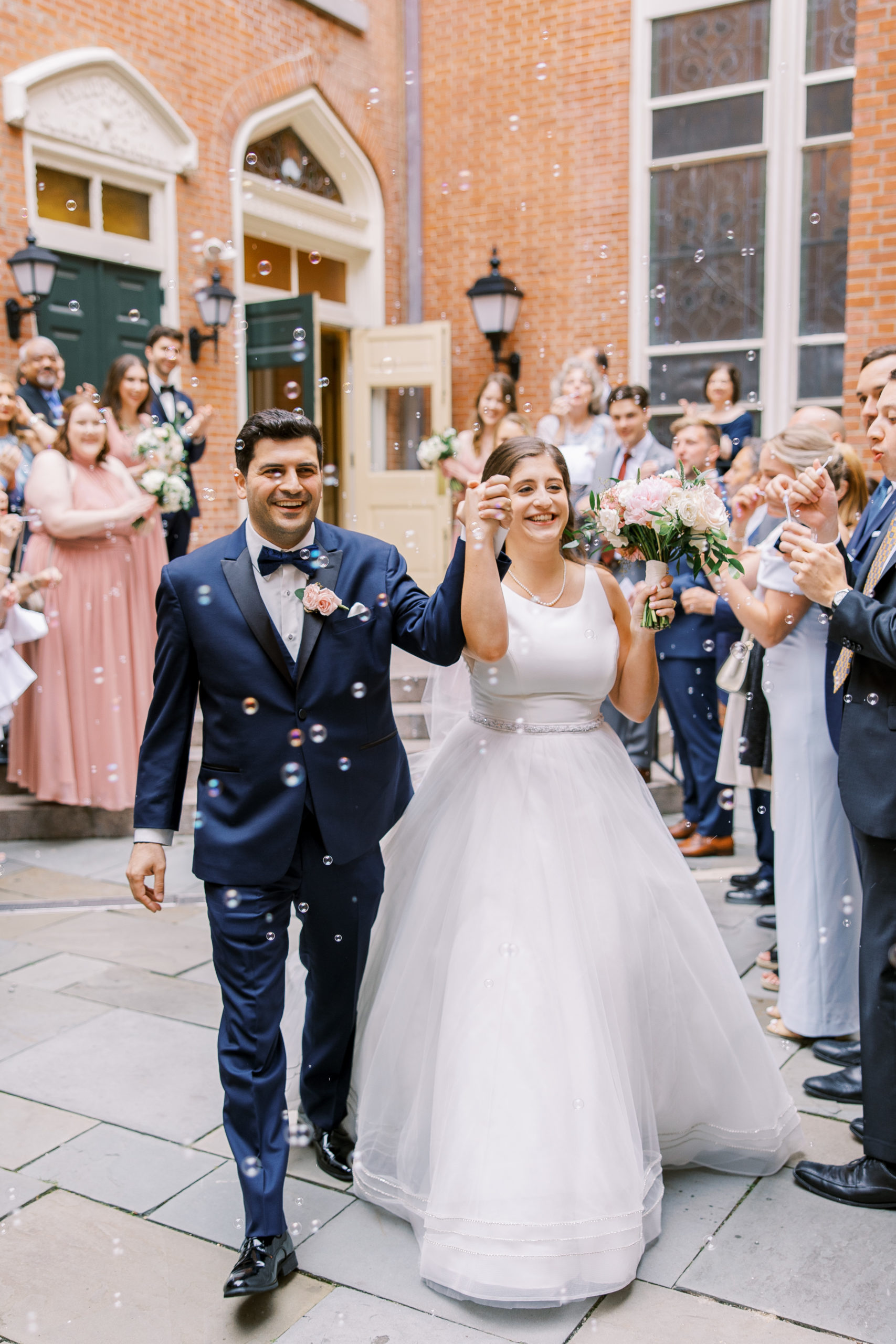 Bride and groom hold hands walking out of church with bubbles around them and friends and family clapping