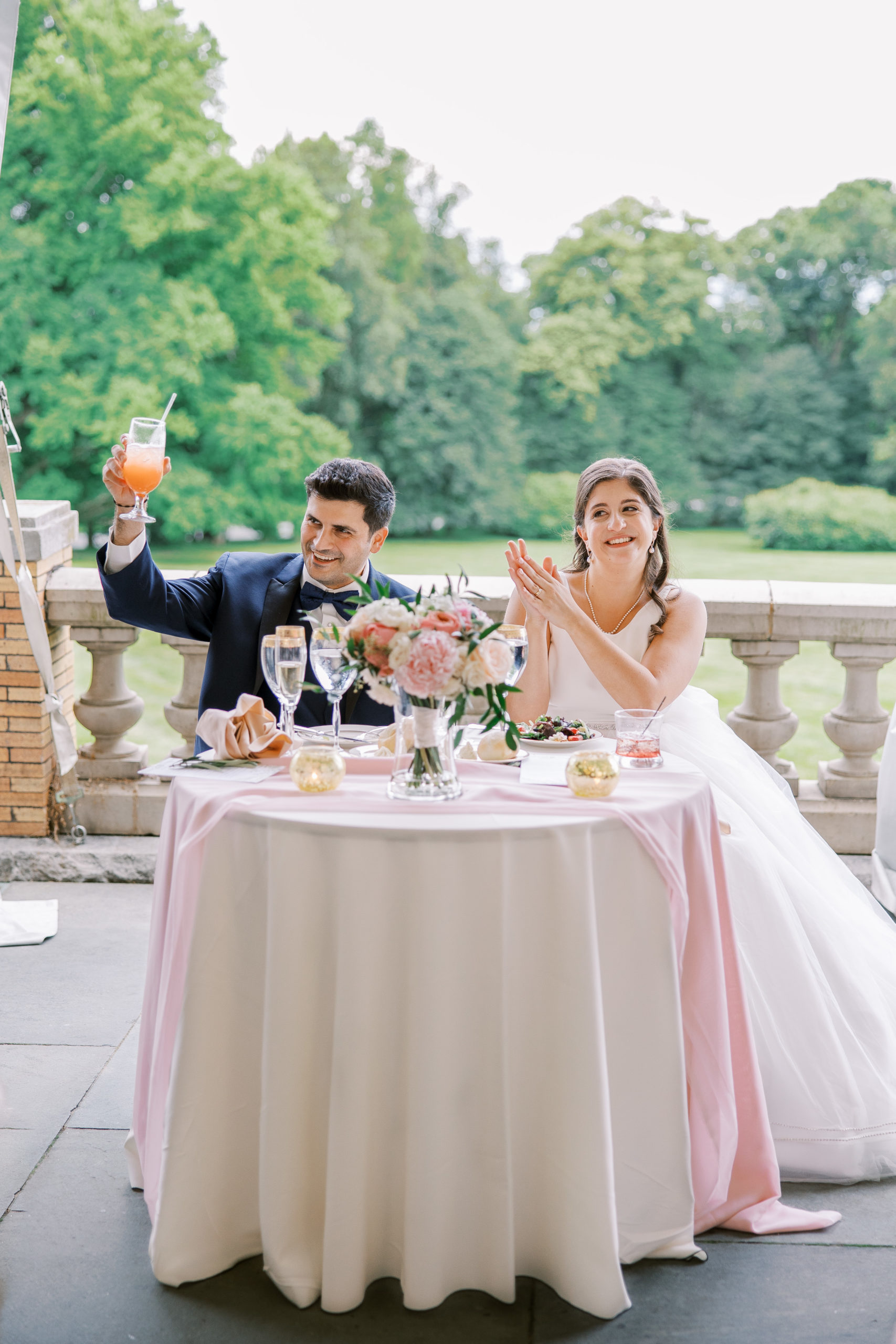 Groom holds up drink and bride claps sitting at their table during wedding reception for Cairnwood Estate Wedding Photography