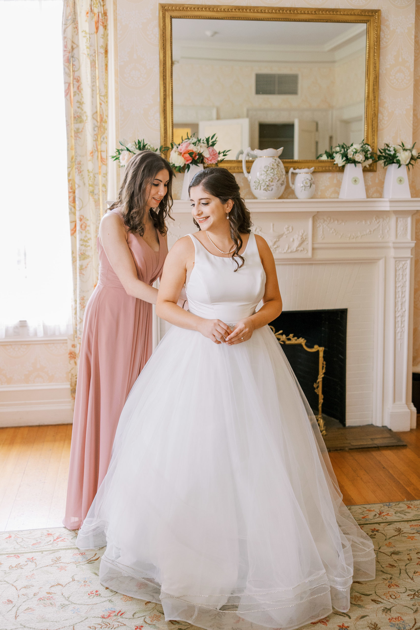 Bridesmaid helps bride with dress next to fireplace and gold mirror at Cairnwood Estate Wedding Photography