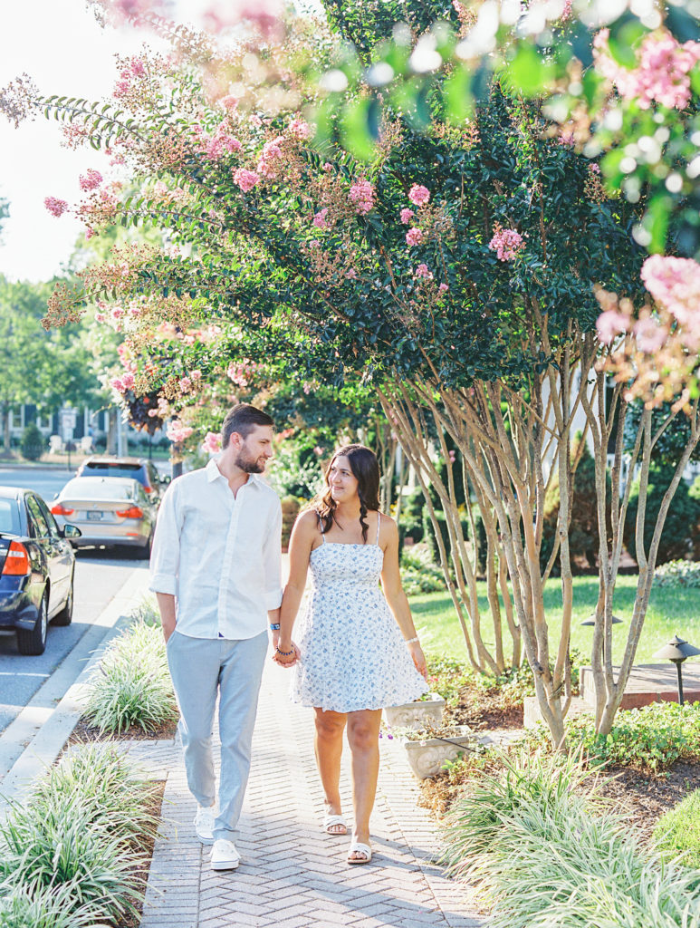 Couple holds hands walking down the street with pink flower tree for Summer Lewes Engagement Session
