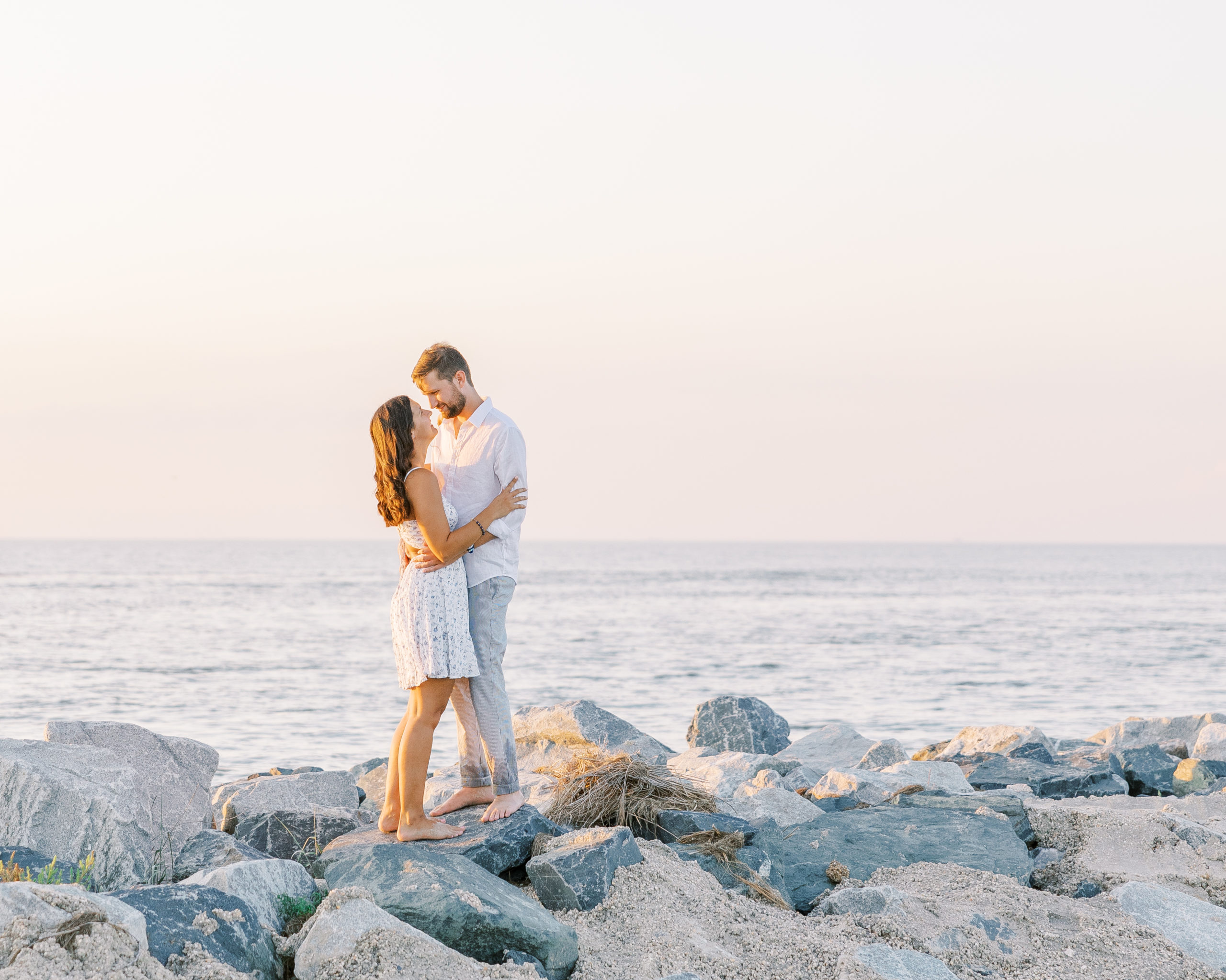 Couple embraces and smiles standing along the jetty overlooking the ocean for Summer Lewes Engagement Session
