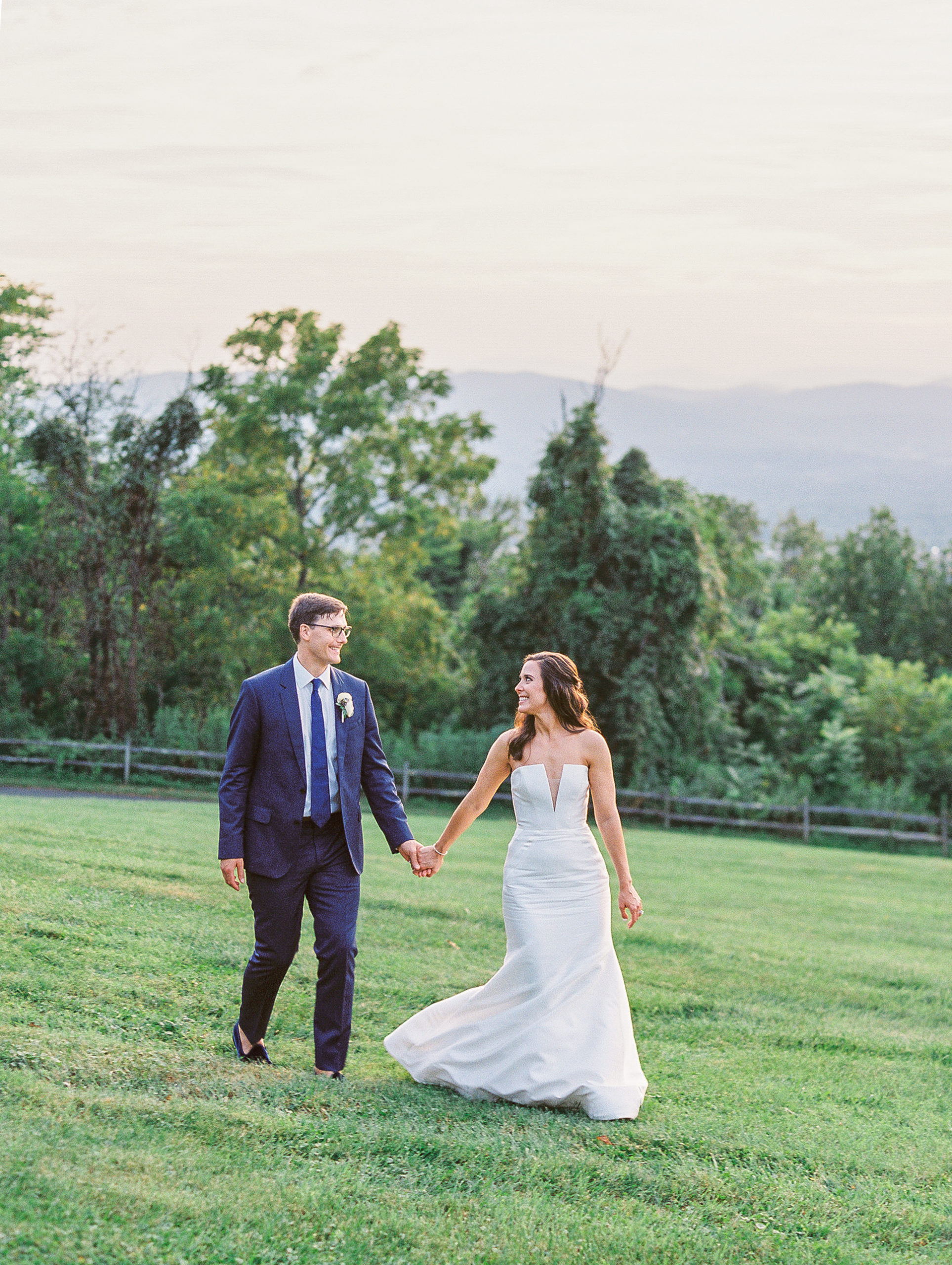 Bride and groom smile holding hands walking in green field on mountain for Charlottesville Wedding Photography
