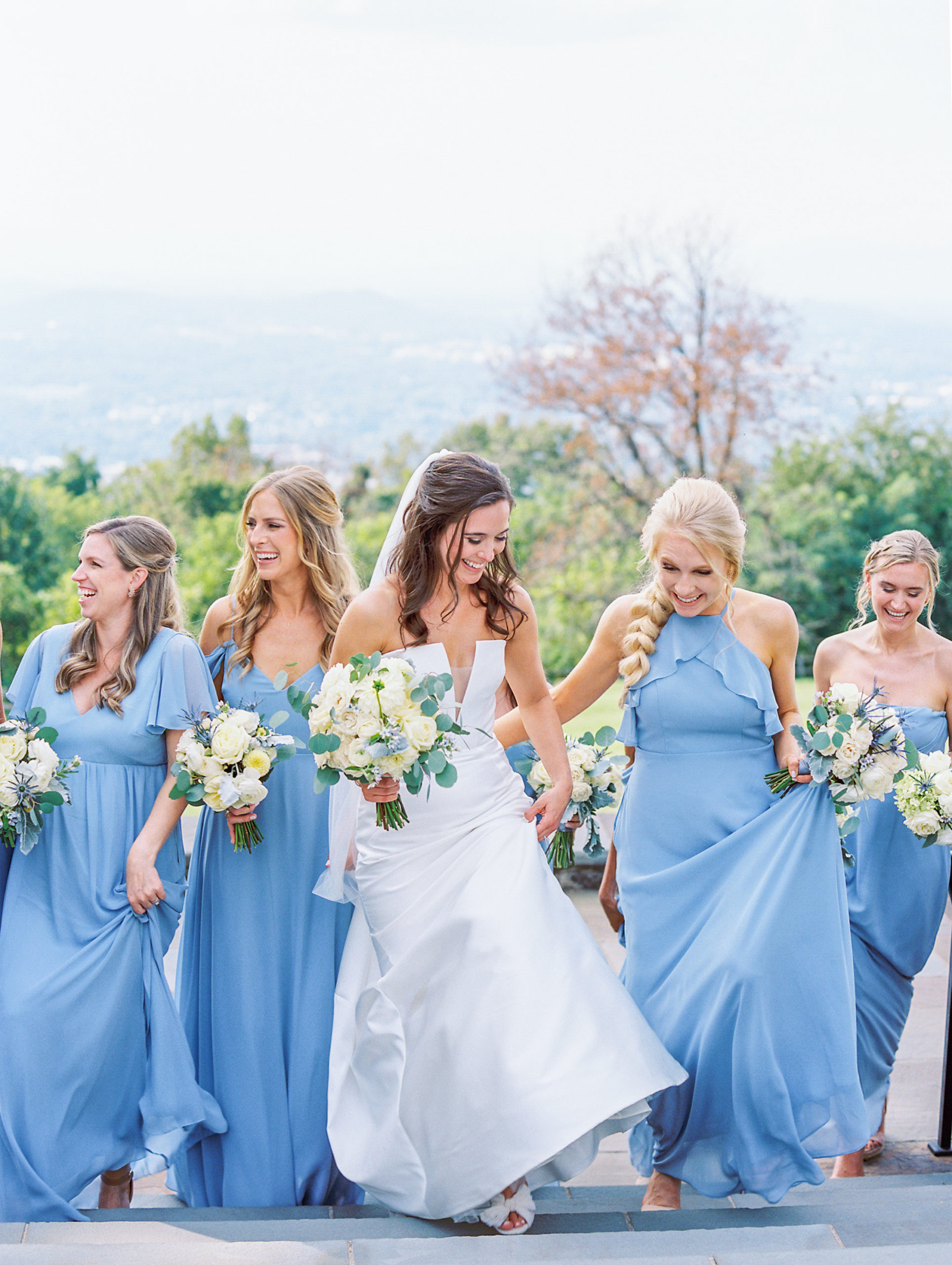 Bridesmaids in blue dresses walk with bride and smile holding rose bouquets 