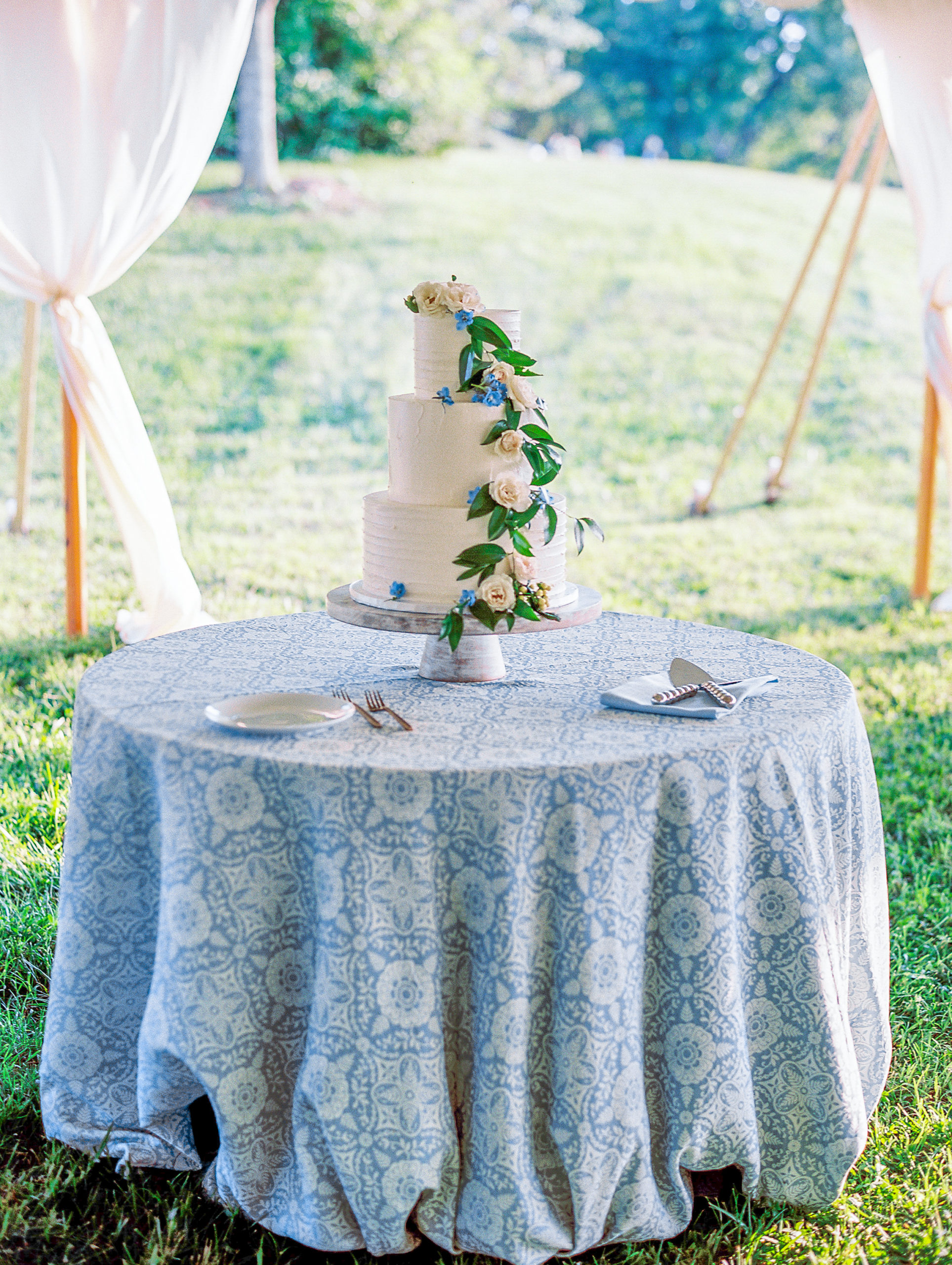 Three tiered white wedding cake with roses and leaves on top of small round and blue table 