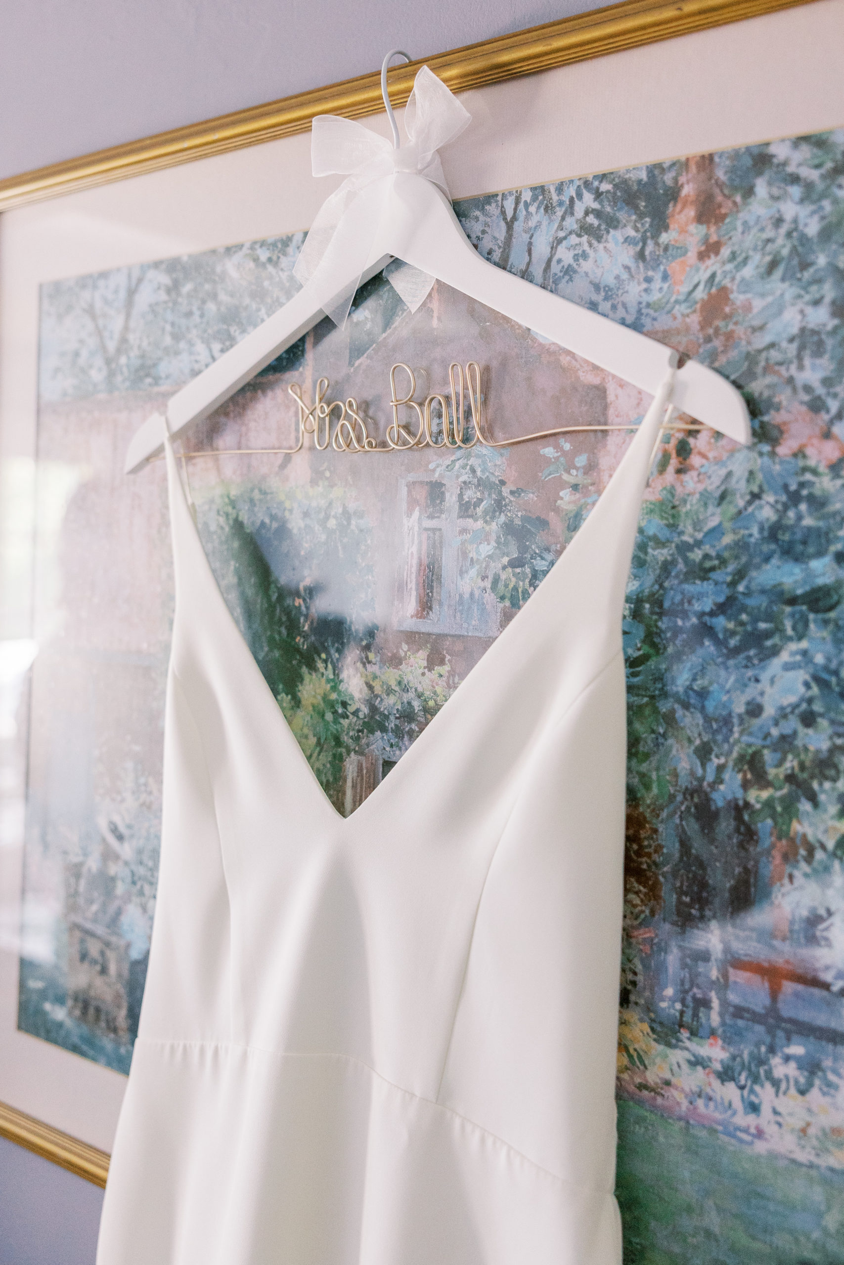 Wedding dress hangs in front of painting with a ribbon and "Miss Ball" in gold attached to hanger 