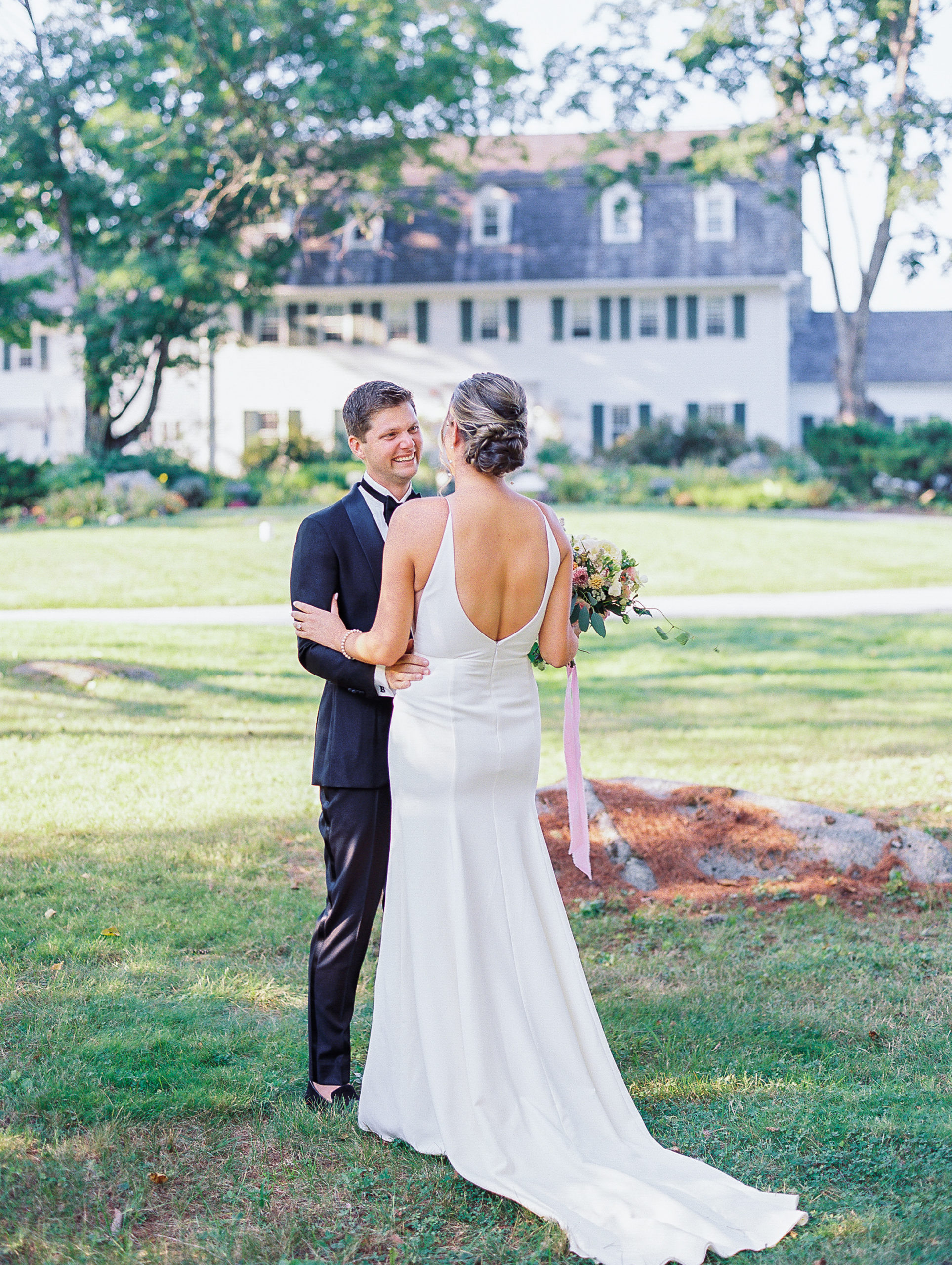 Bride and groom smile and embrace in front of white paneled house 