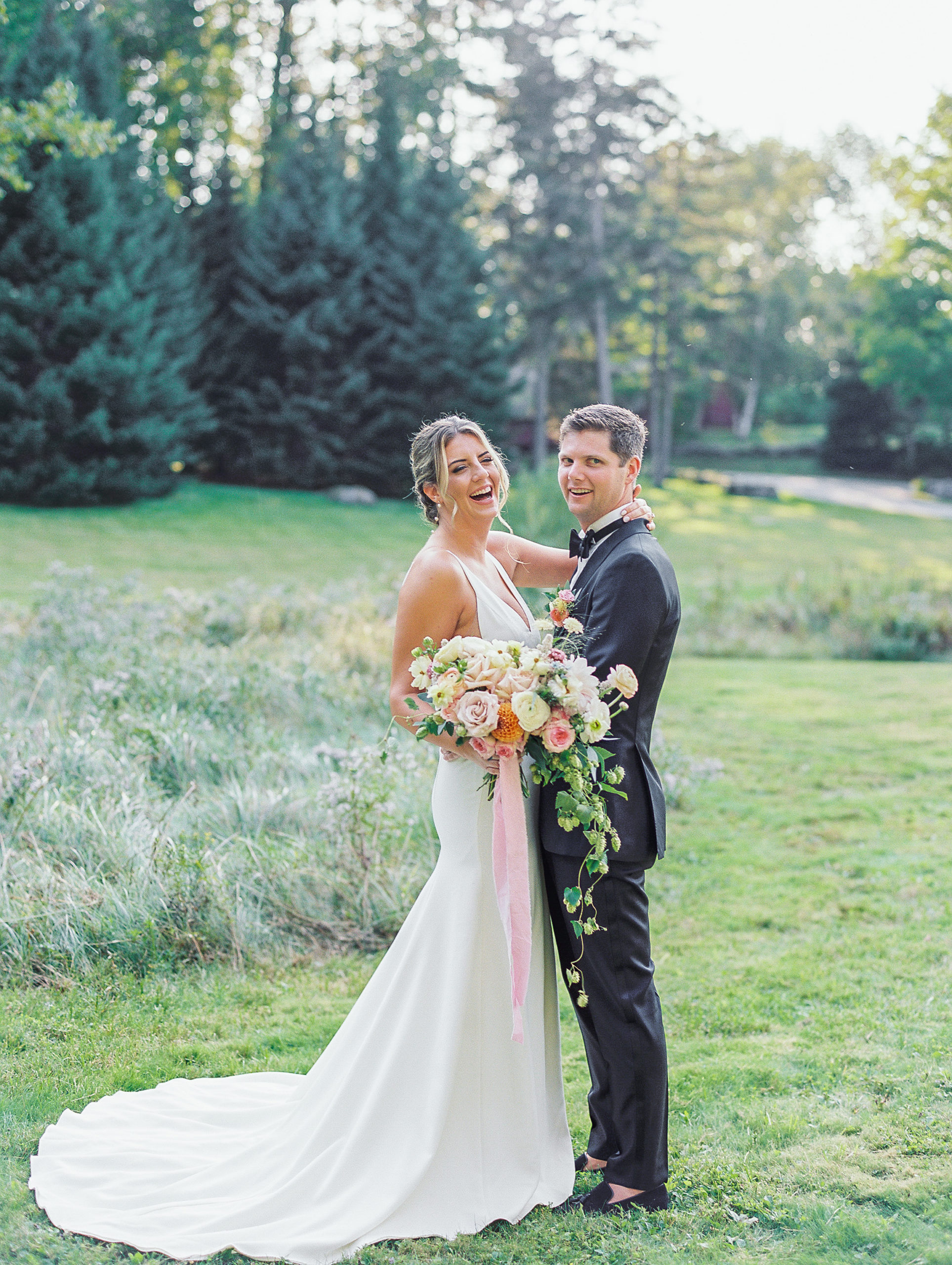 Bride laughs and holds bouquet and groom laughs smiles in open green landscape for New Hampshire Wedding Photography