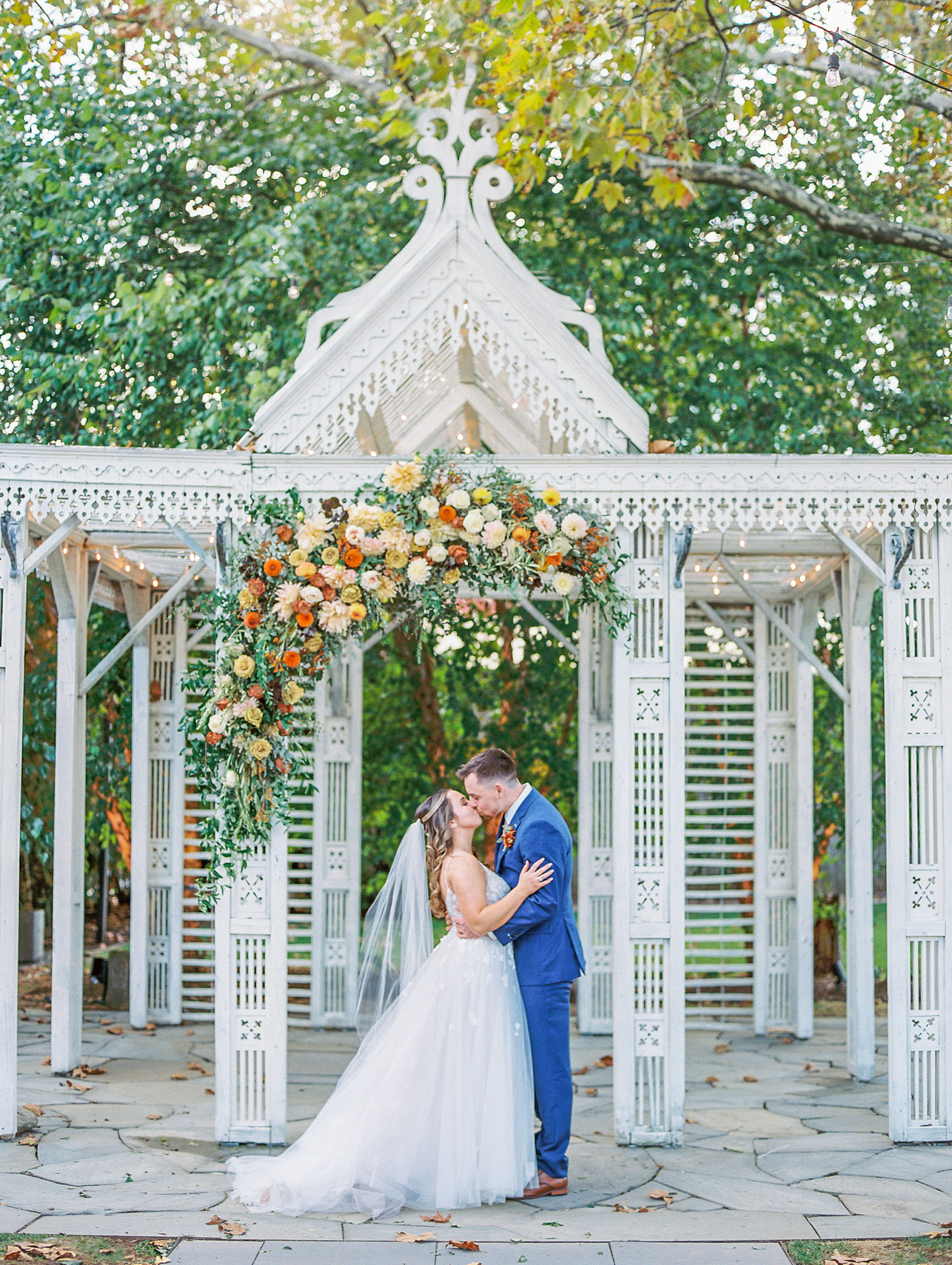 Bride and groom embrace and kiss in front of white pavilion with beautiful arrangement of autumnal colored flowers above 