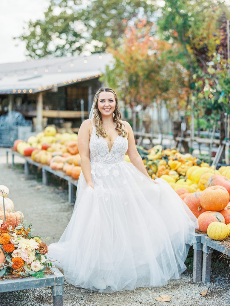 Bride holds dress and smiles next to assortment of flowers and pumpkins at terrain 