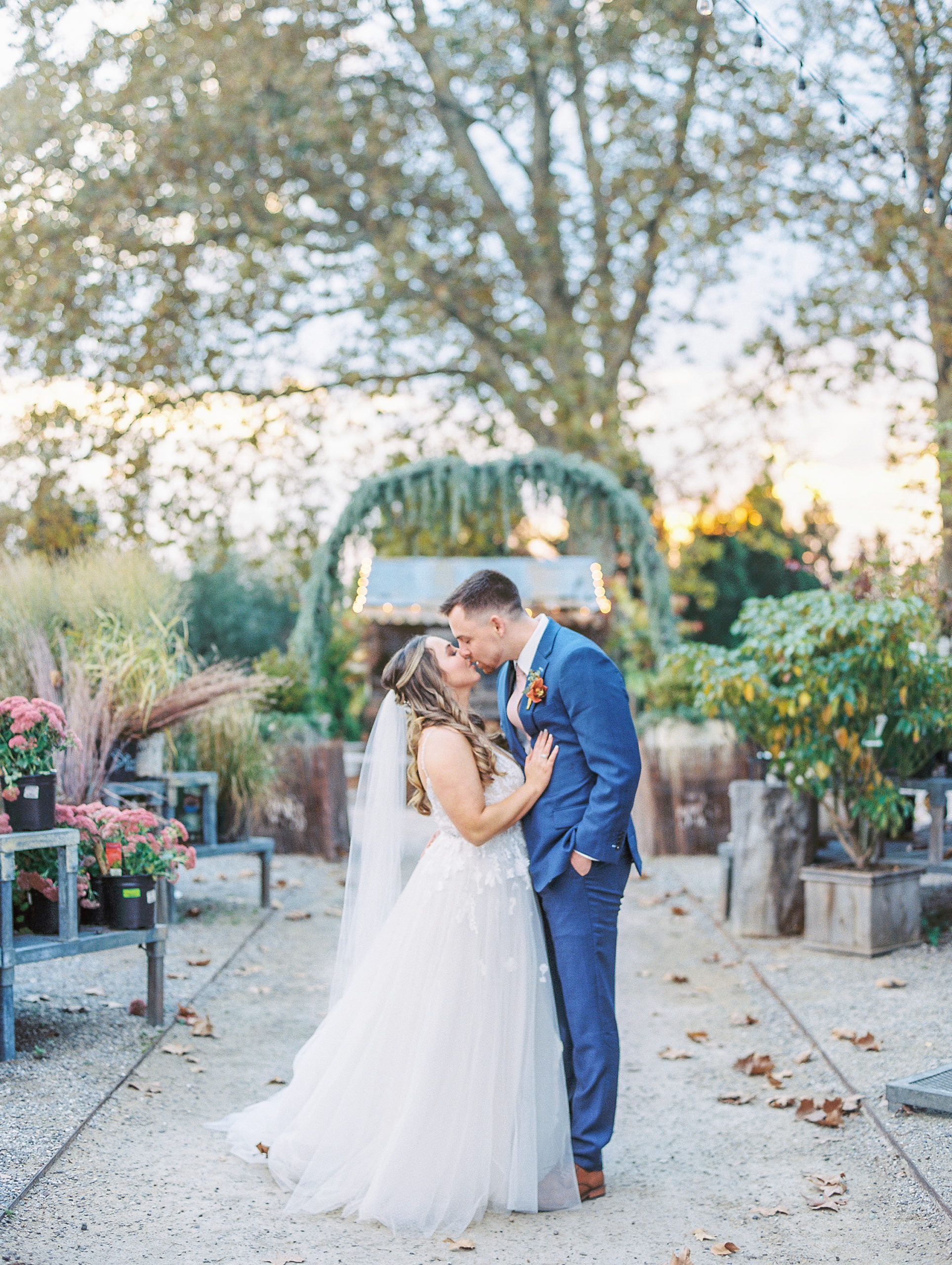 Bride and groom share a kiss along gravel pathway with greenery under arch for Terrain Glen Mills Wedding
