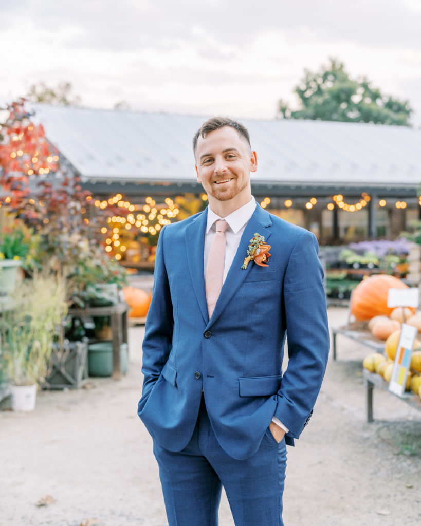 Grooms poses with hands in pocket with blue suit and light pink tie at terrain gardens 